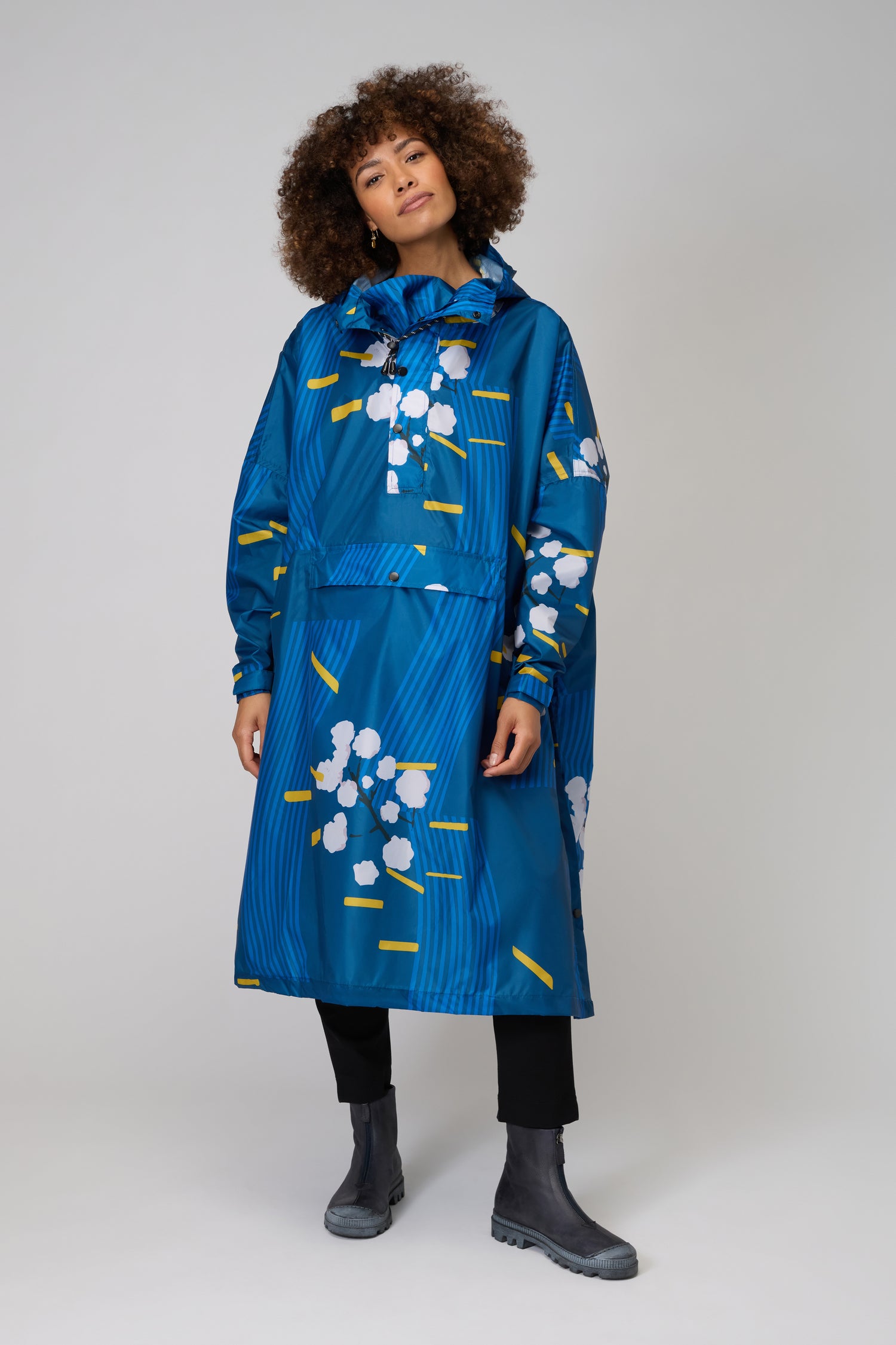 A person wears the Japanese Blue Blossom Poncho—made from recycled plastic—with white and yellow abstract prints over blue, paired with black pants and gray boots, standing against a plain light background. Stylish and sustainable.