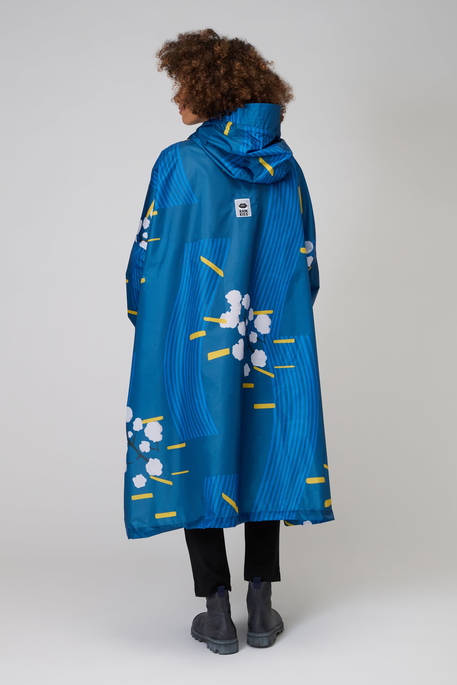 A person faces away against a plain background, wearing the Japanese Blue Blossom Poncho—waterproof, made from recycled plastic, with a blue hood and abstract yellow, white, and blue patterns.