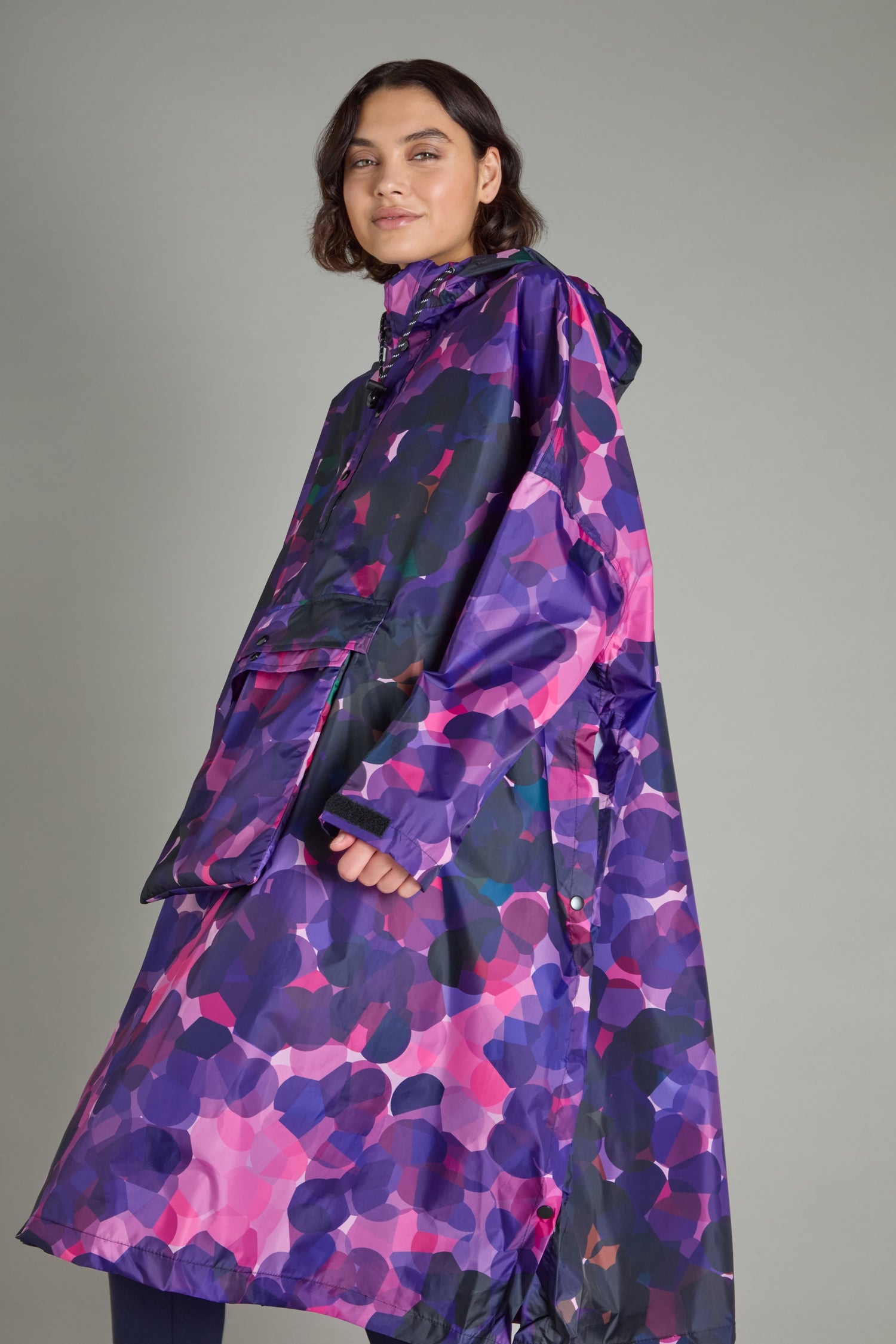 A person wearing the City Lights Poncho, featuring a purple and pink camouflage pattern and made from recycled plastic, stands against a gray background.