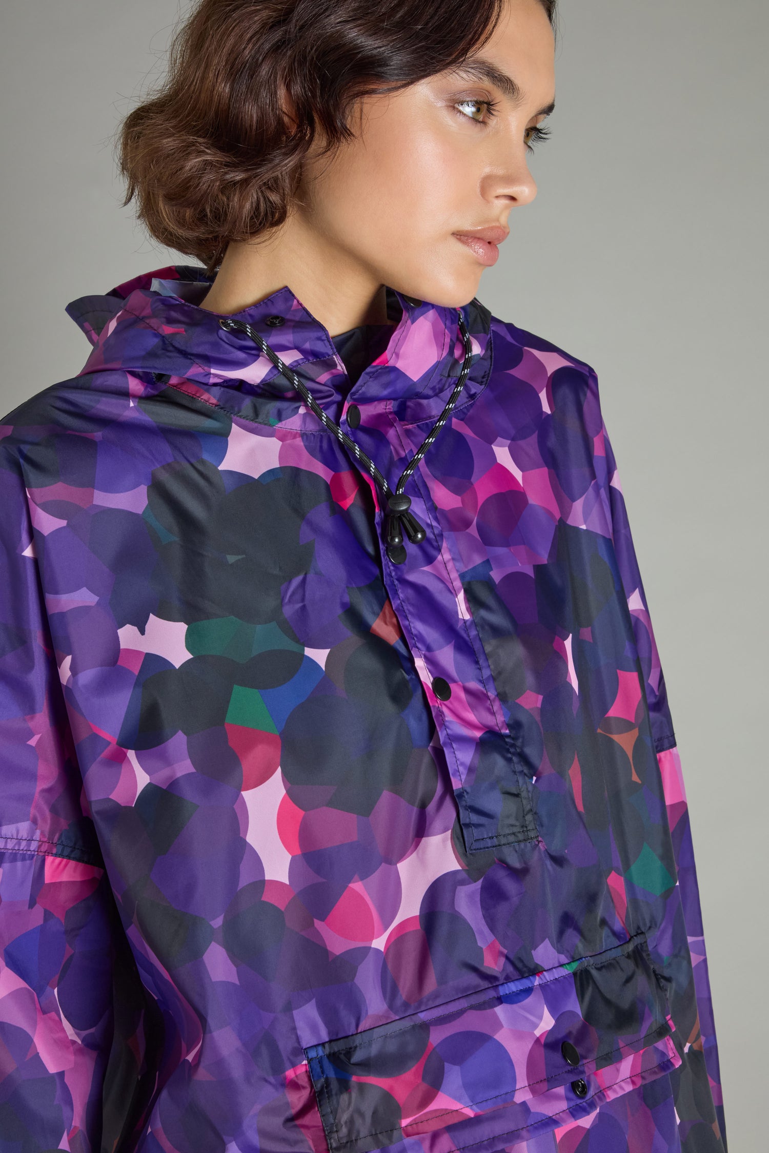 A person wearing the City Lights Poncho, featuring a purple and pink pattern and made from recycled plastic, looking to the side.