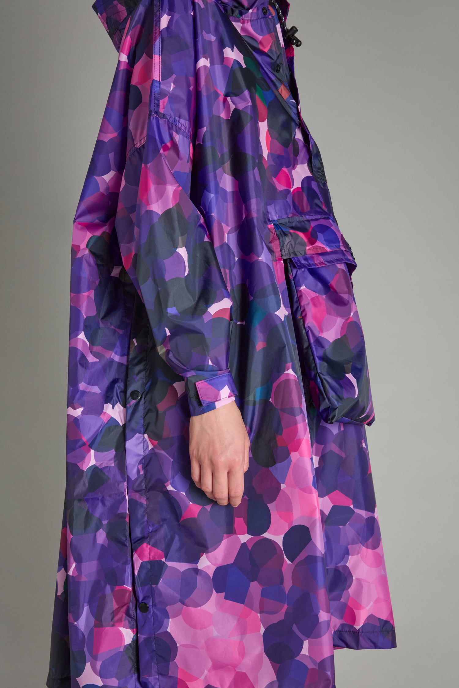 A person wearing the City Lights Poncho, a raincoat made from recycled plastic with a purple and pink camouflage pattern and featuring a large front pocket.
