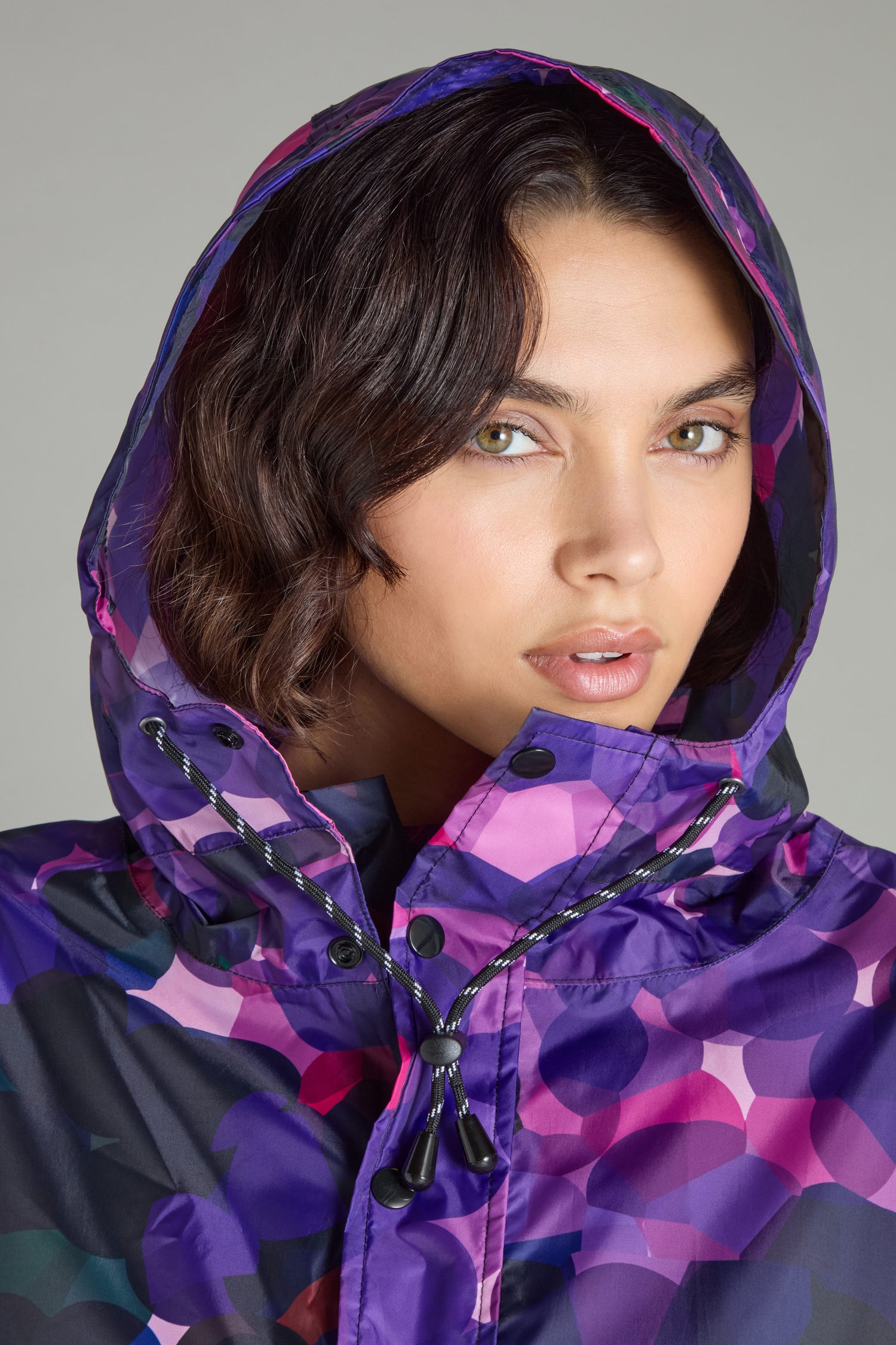 A person is wearing the City Lights Poncho, a purple and pink patterned hooded jacket made from recycled plastic, gazing intently at the camera.