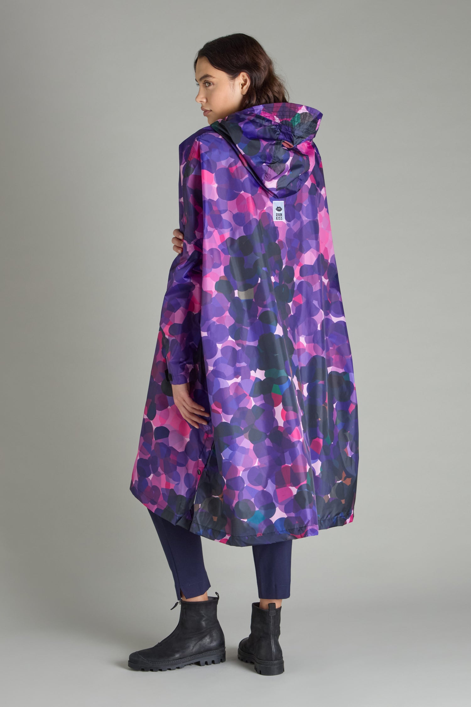 A person stands against a plain gray backdrop, wearing the City Lights Poncho—a colorful, patterned raincoat crafted from recycled plastic. The hooded design pairs perfectly with black pants and boots, seamlessly blending sustainability with style in an urban aesthetic.