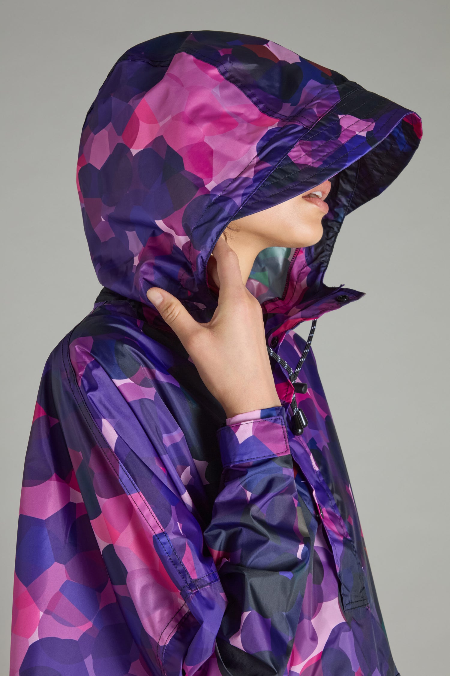 A person wearing the City Lights Poncho, made from recycled plastic, displays a purple and pink geometric pattern. They face sideways with their hand near the neck, highlighting its urban design flair.