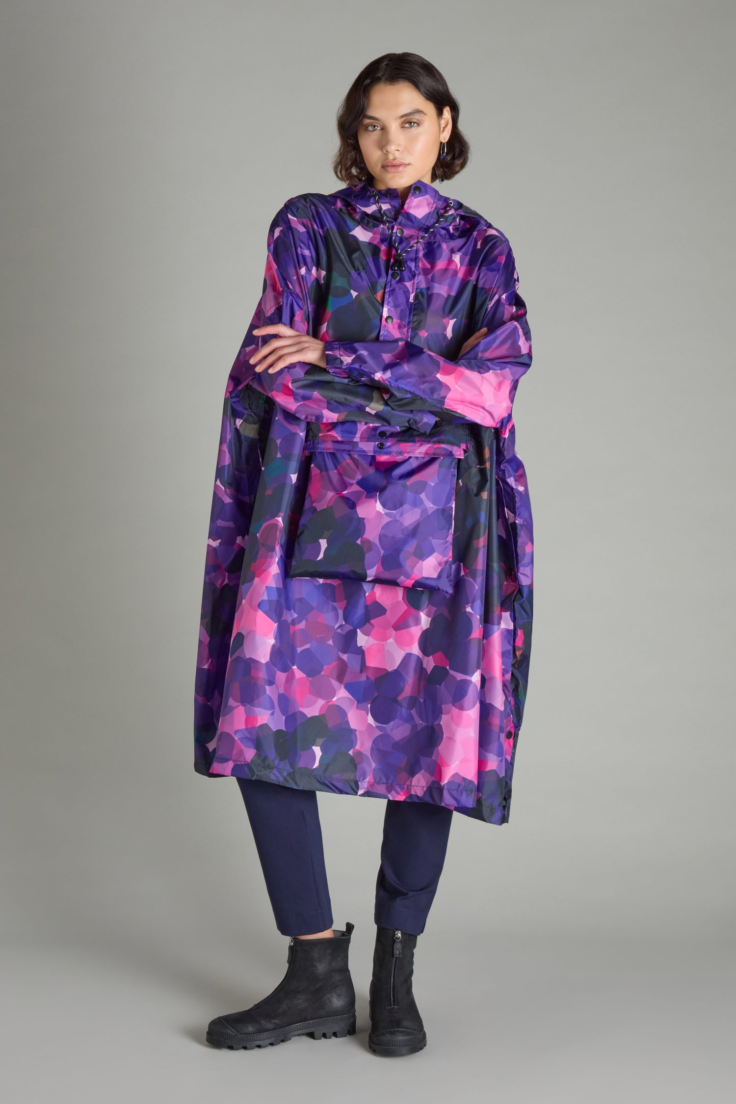 A person wearing the City Lights Poncho, a colorful and patterned waterproof garment made from recycled plastic, along with dark pants and black boots, stands against a plain background.