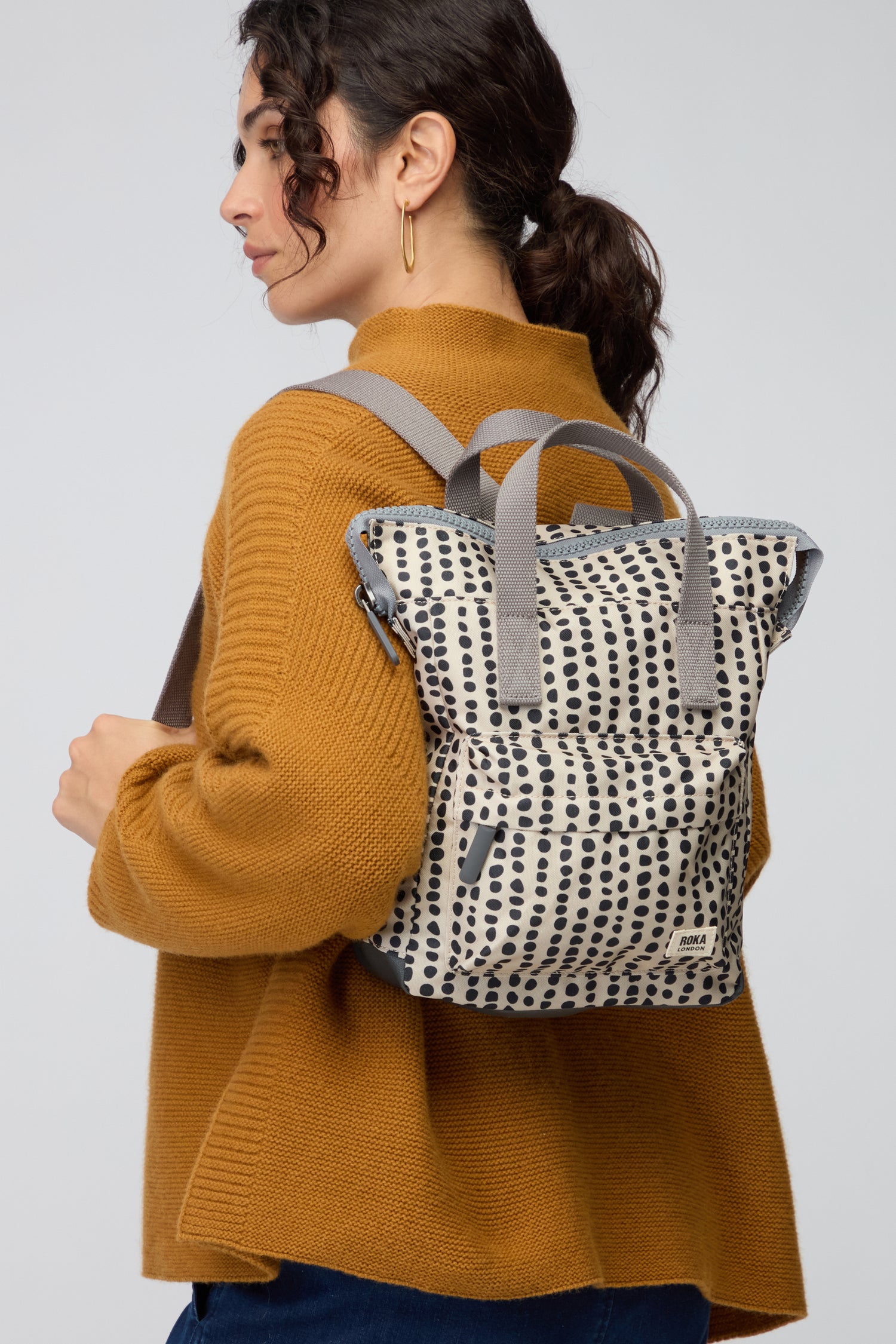 A woman wears a mustard sweater and carries the Recycled Canvas Bantry Dotty Bag—a small white backpack with black polka dots and gray straps—shown from the side against a neutral background.