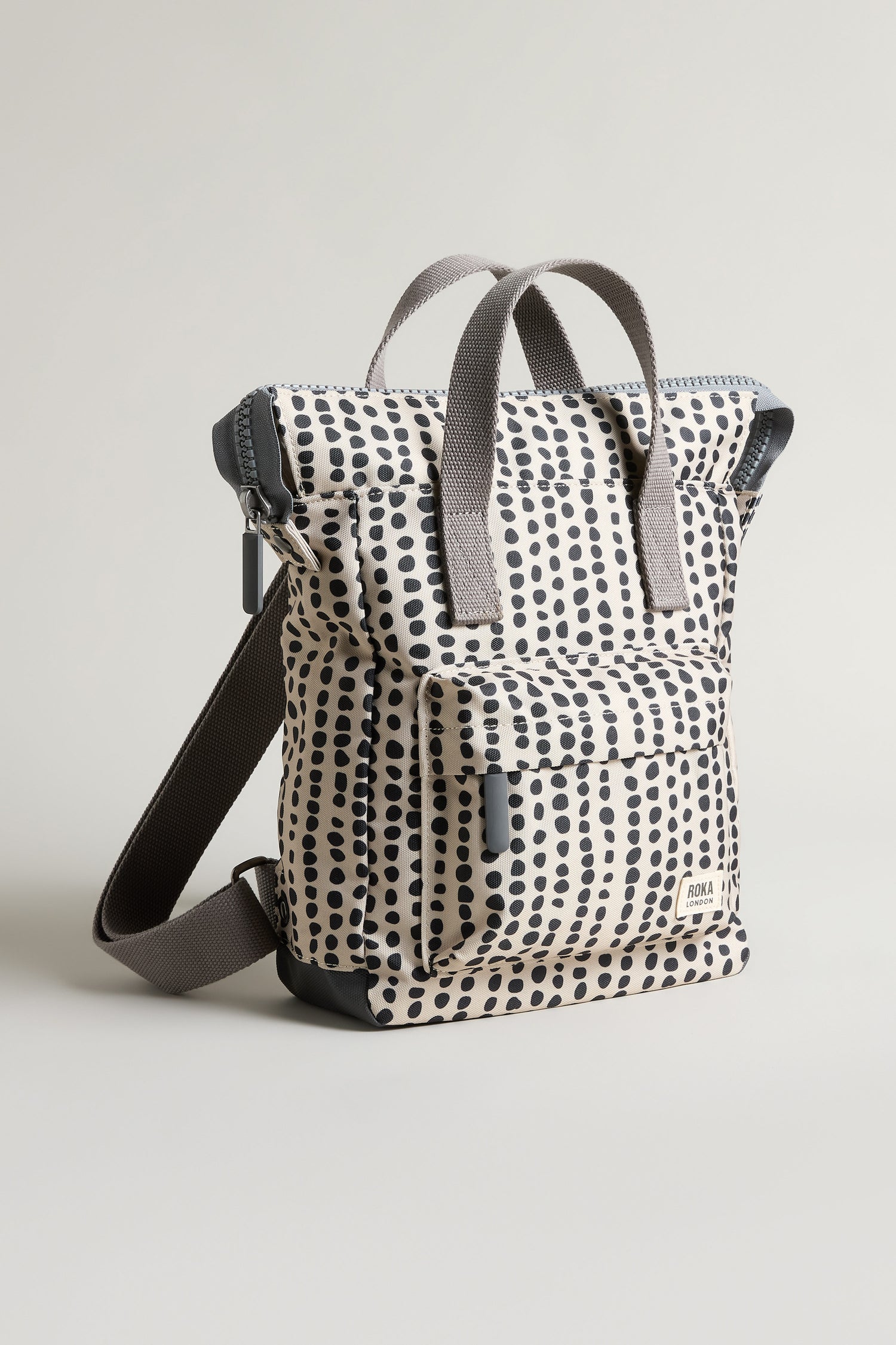 The Recycled Canvas Bantry Dotty Bag is a sustainable cream backpack with black dotted vertical lines, gray handles, a front pocket, and an adjustable dark shoulder strap, shown against a plain light background.
