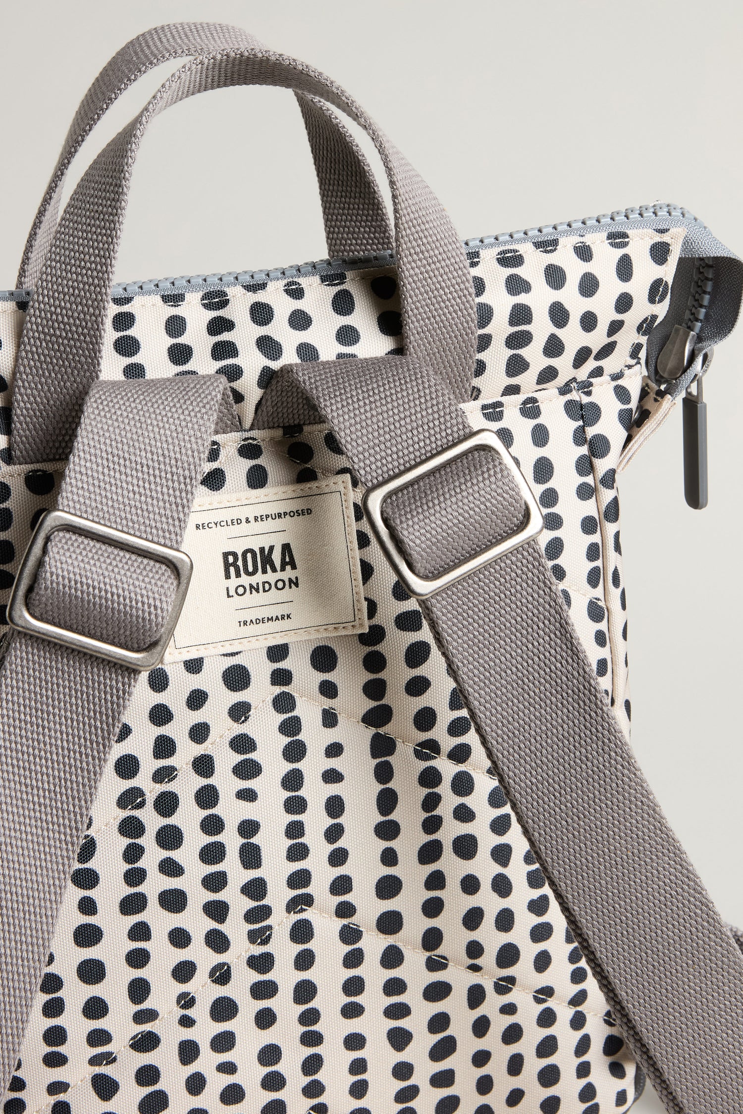 Close-up of the Recycled Canvas Bantry Dotty Bag, a weather-resistant white backpack with black polka dots, grey straps, and a visible "ROKA London" label on the back.
