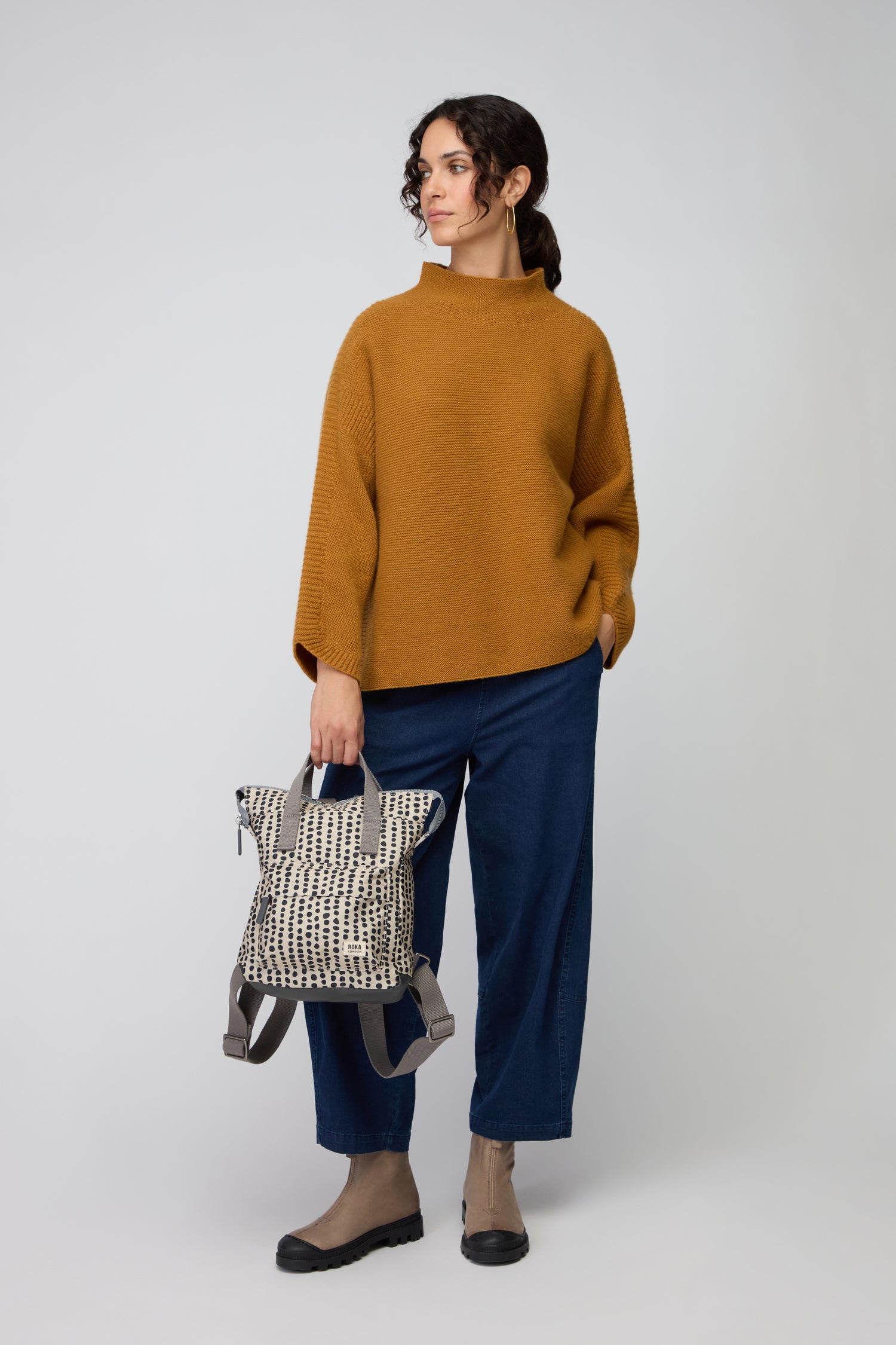 A person stands against a plain background in a mustard sweater, blue jeans, taupe boots, and holds the Recycled Canvas Bantry Dotty Bag.