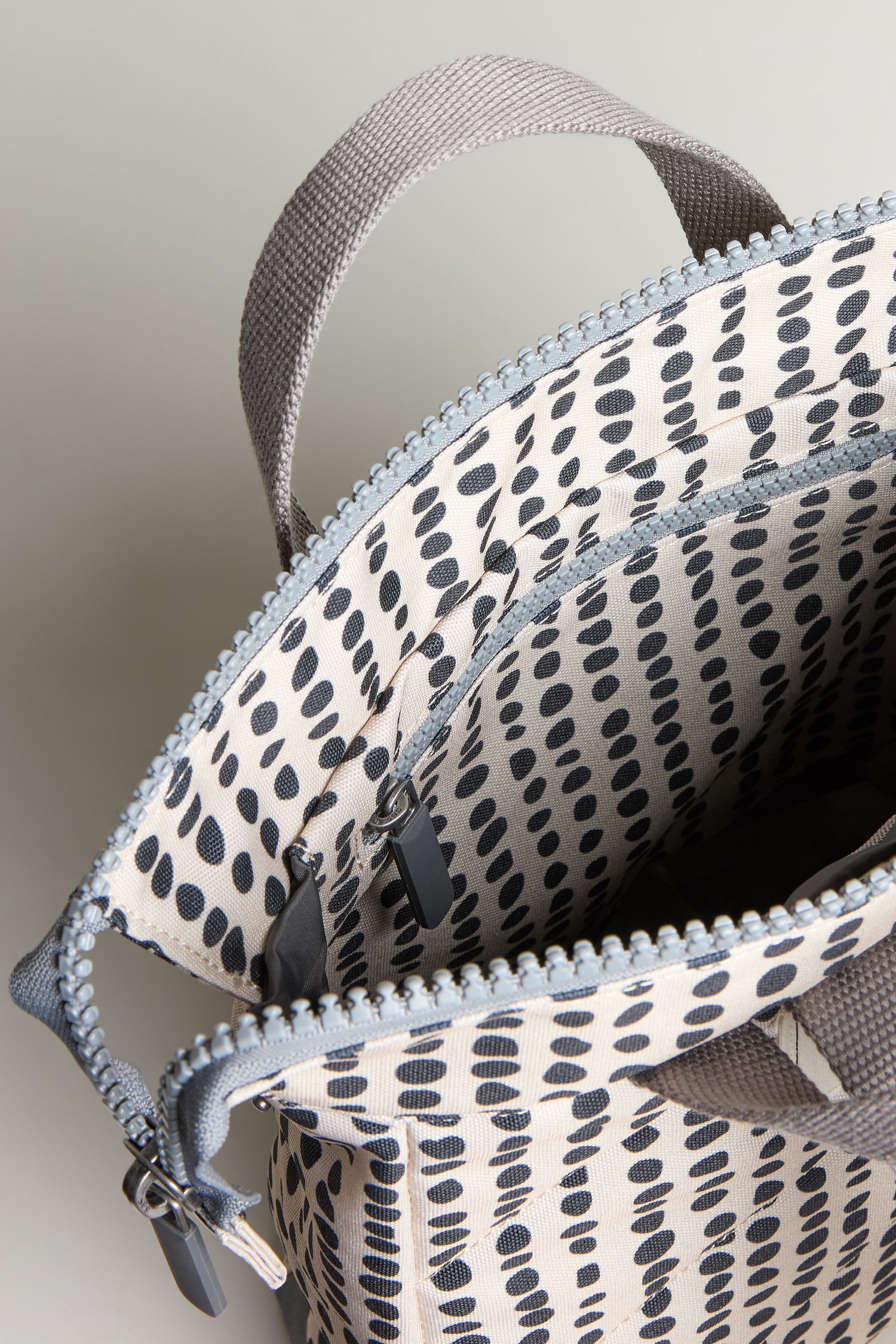 A close-up of the Recycled Canvas Bantry Dotty Bag, open to show its inner pocket, with a grey zipper and handle, and a black dotted pattern on a white background.