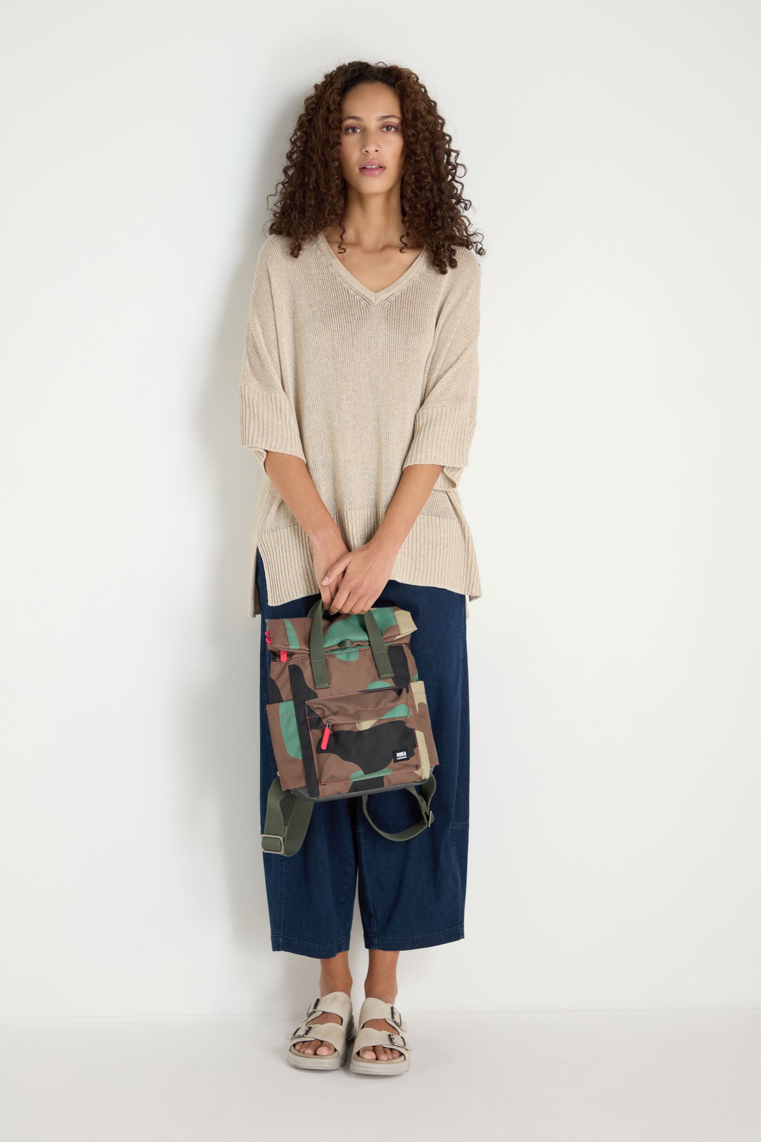 Woman with curly hair stands against a white wall, wearing a beige sweater, blue wide-leg pants, white sandals, and holding the Recycled Canvas Canfield Bag in front of her.