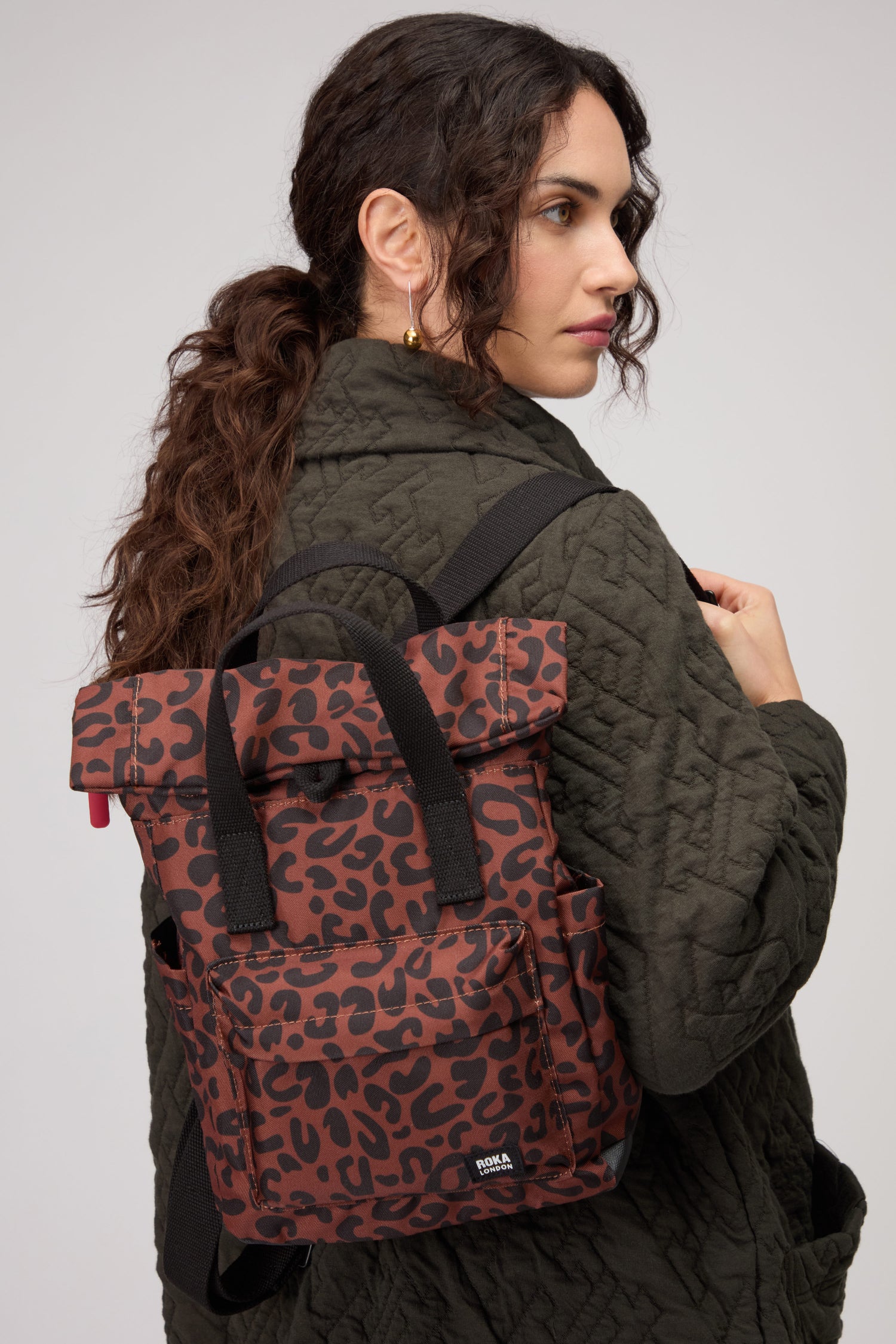 A person wearing an olive quilted jacket carries the Recycled Canvas Canfield Leopard Backpack in bold red and black leopard print, glancing over their shoulder against a plain background.