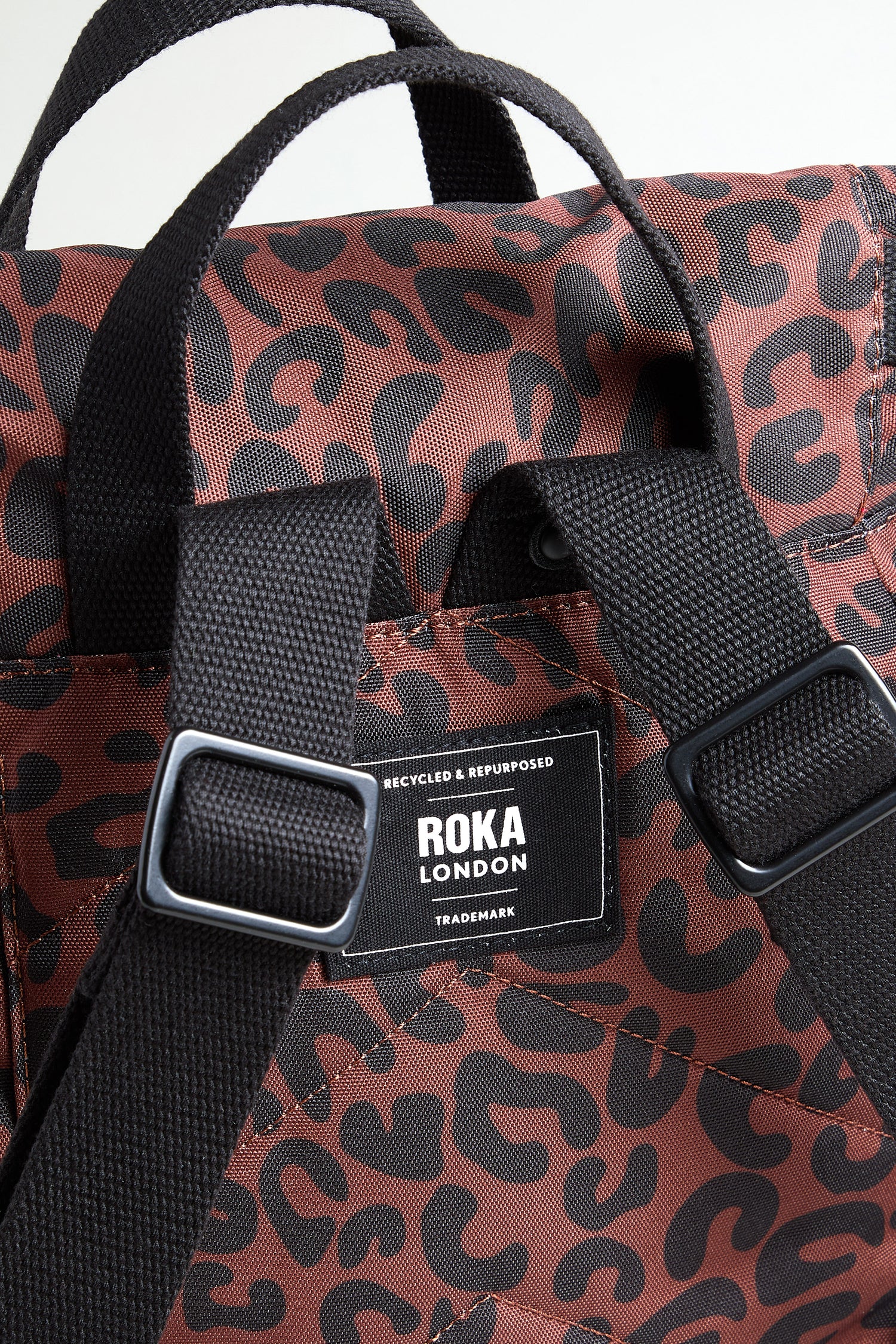 Close-up of the Recycled Canvas Canfield Leopard Backpack, featuring brown and black leopard print, black straps, water-resistant recycled canvas, and a "Roka London" label on the back.