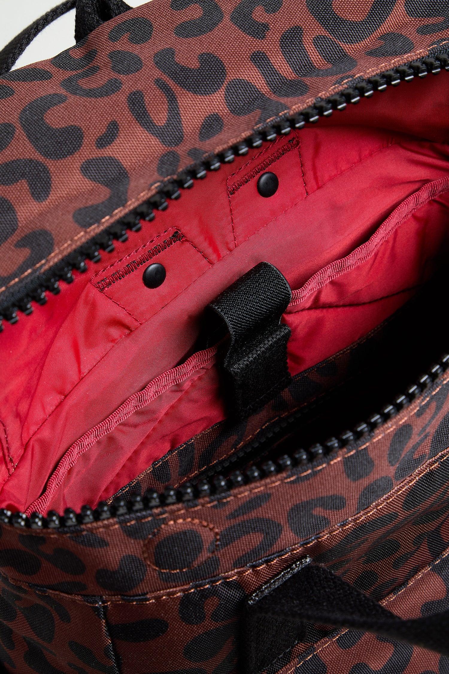 Close-up of the Recycled Canvas Canfield Leopard Backpack featuring a brown and black leopard print exterior, red lining, interior pocket, black strap, and durable water-resistant recycled canvas for style and function.