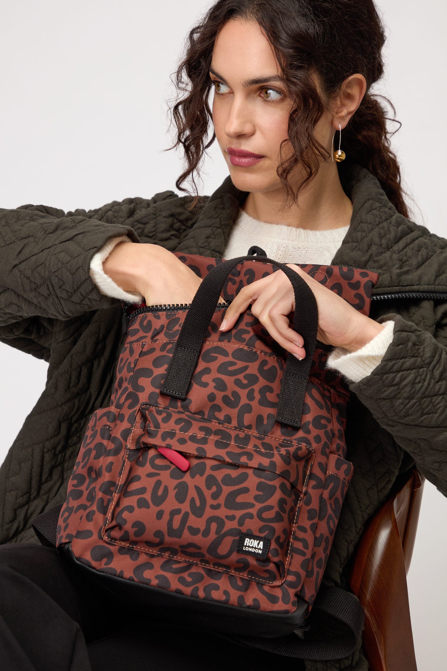 A woman reaches into the Recycled Canvas Canfield Leopard Backpack, a stylish, water-resistant bag featuring a chic brown and black leopard print, paired with her quilted jacket and cream sweater.