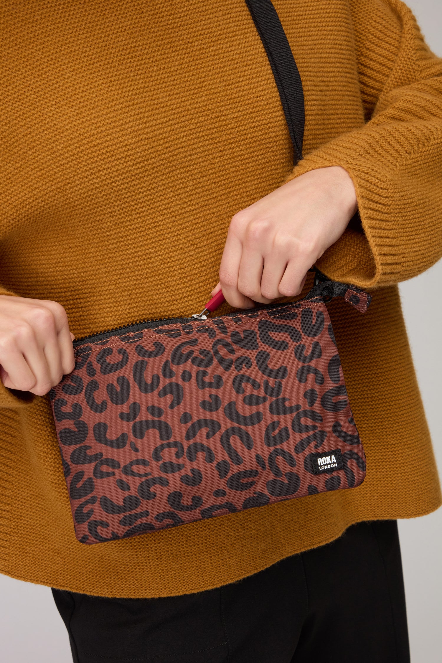 A person in a mustard sweater unzips the Recycled Canvas Carnaby Leopard Crossbody Bag by ROKA, wearing its black shoulder strap. Made from recycled canvas with a bold leopard print, this stylish bag is perfect for everyday use.