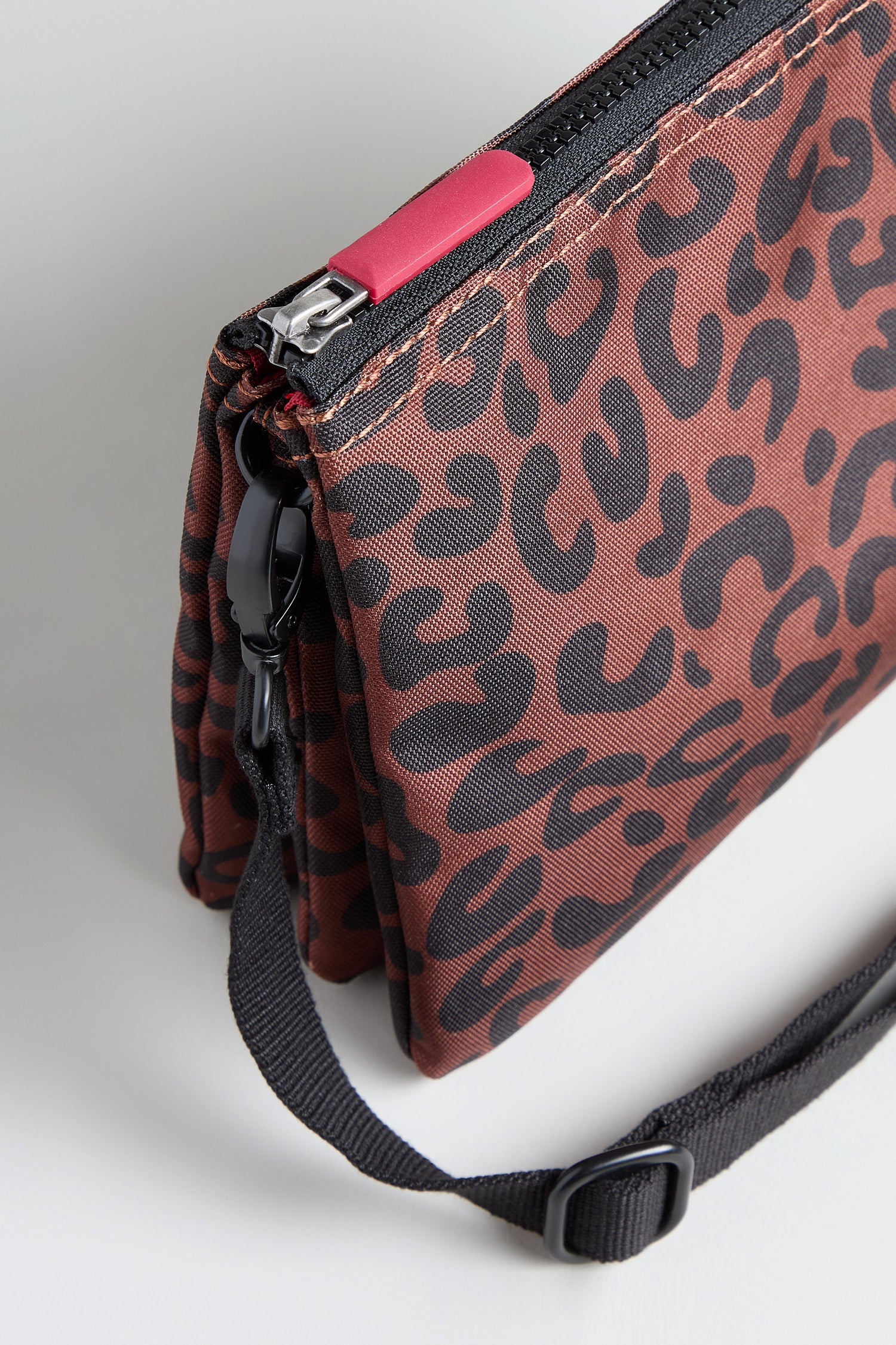 A close-up of the Recycled Canvas Carnaby Leopard Crossbody Bag, showcasing its black and brown leopard print, black adjustable strap, and a zipper with a red pull tab.