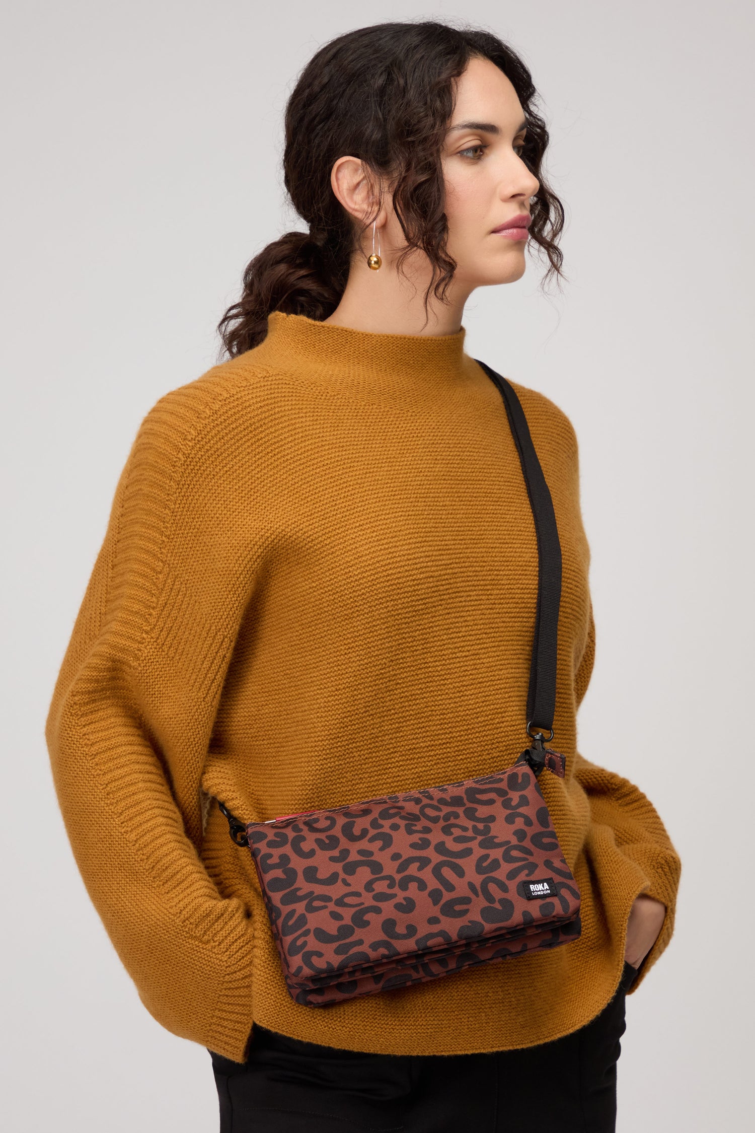 A person in a mustard sweater and black pants stands with hands in pockets, carrying the Recycled Canvas Carnaby Leopard Crossbody Bag across their shoulder.