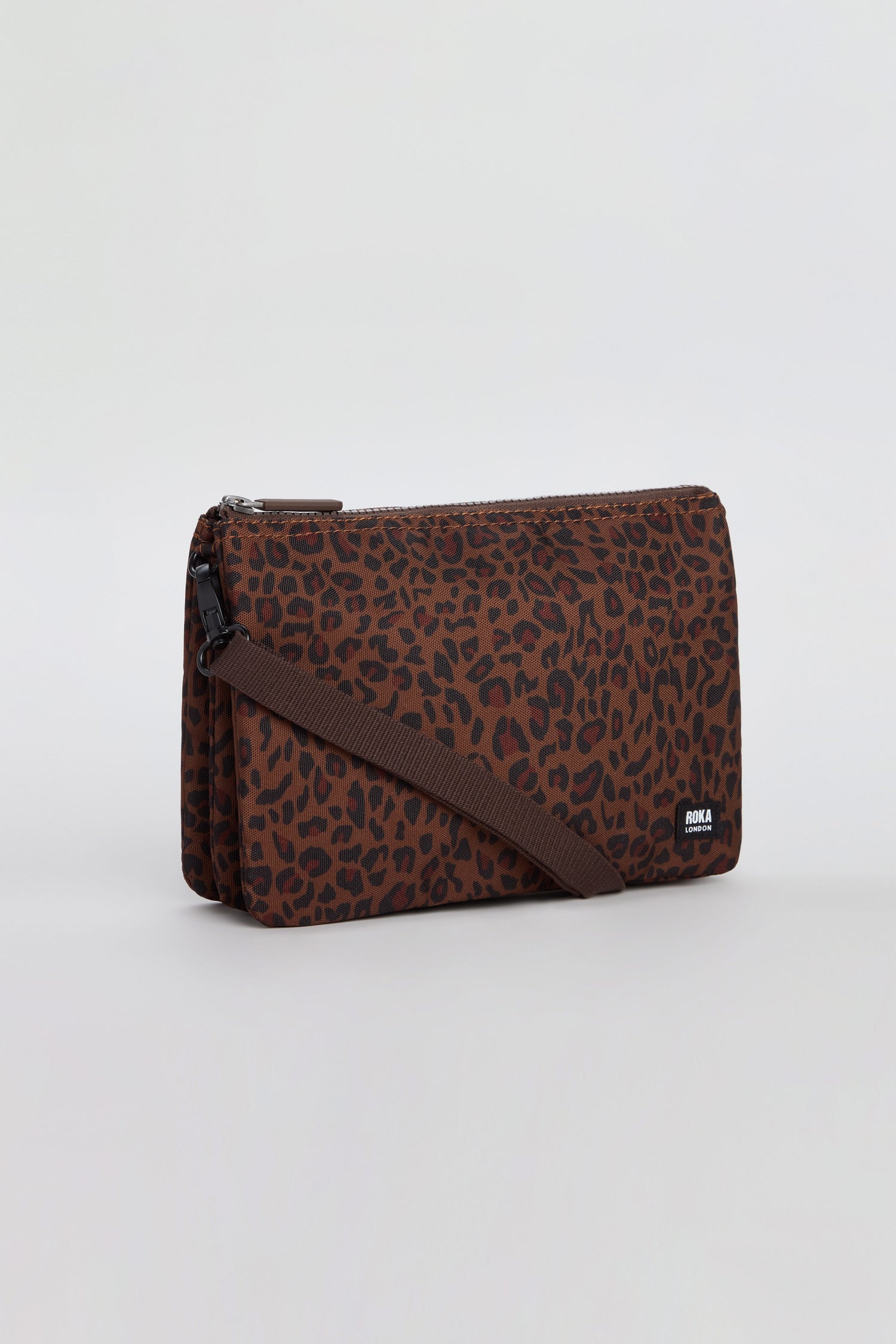 The Mini Leopard Carnaby Crossbody Canvas Bag features a brown rectangular shape with a bold black and dark red leopard print pattern, a zipper closure, and an adjustable brown strap. Crafted from recycled canvas materials, this bag is displayed against a plain light background.