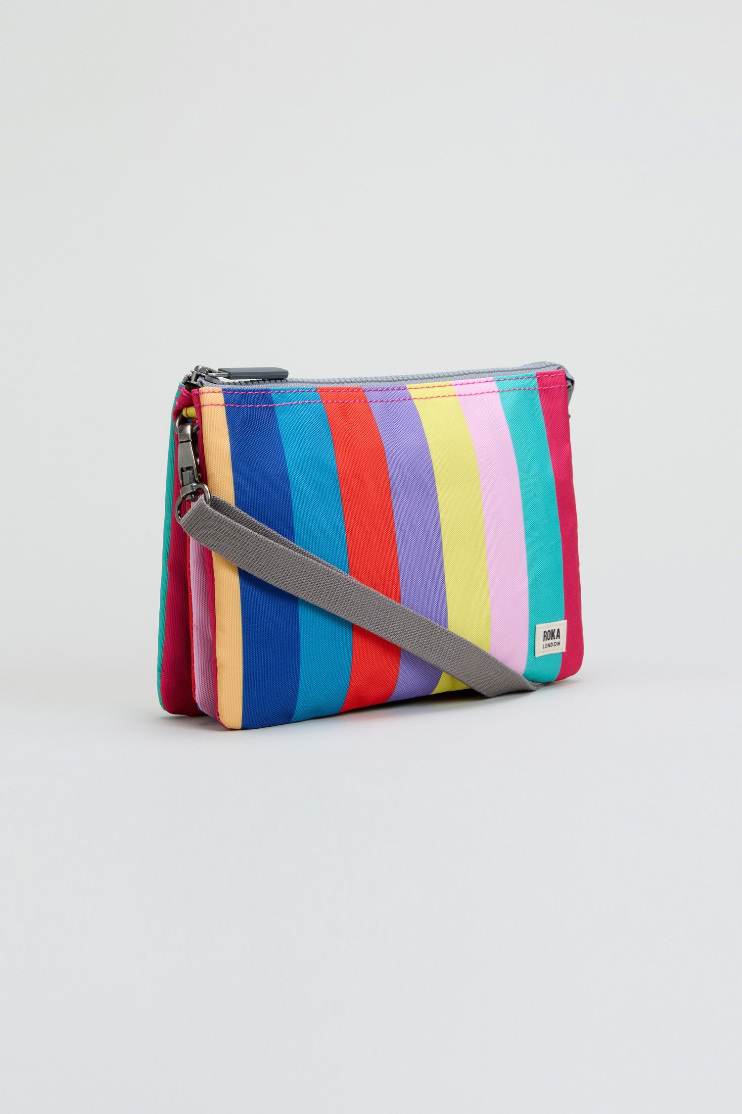 Introducing the Wide Multistripe Carnaby Crossbody Canvas Bag—crafted from recycled canvas, this small bag features bold vertical rainbow-colored stripes and comes with a detachable gray strap, showcased against a simple light background.