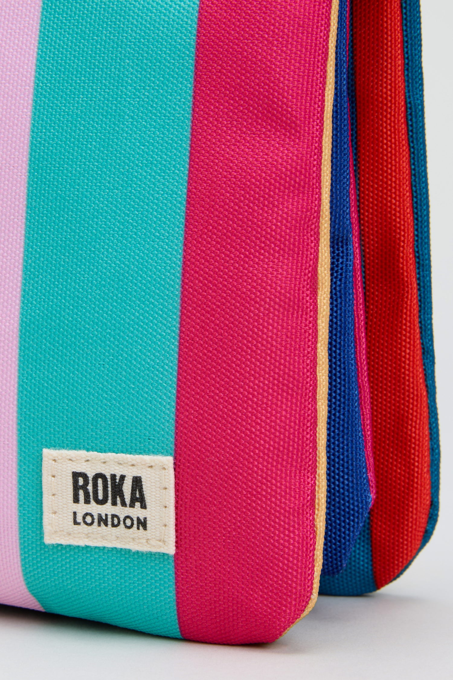A close-up of the Wide Multistripe Carnaby Crossbody Canvas Bag showcases its vibrant pink, teal, blue, and red stripes. Featuring the ROKA London label, this bag is crafted from weather-resistant recycled canvas for both style and durability.