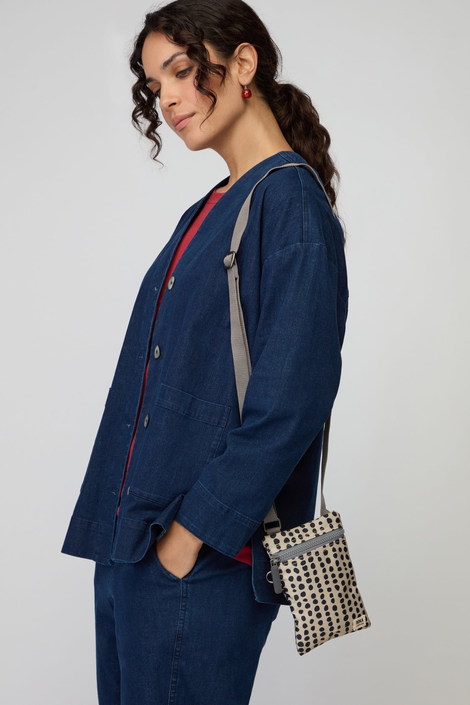 Woman wearing a denim jacket and pants with a red top carries a Recycled Canvas Chelsea Dotty Bag, stands with her hand in her pocket, and looks down.