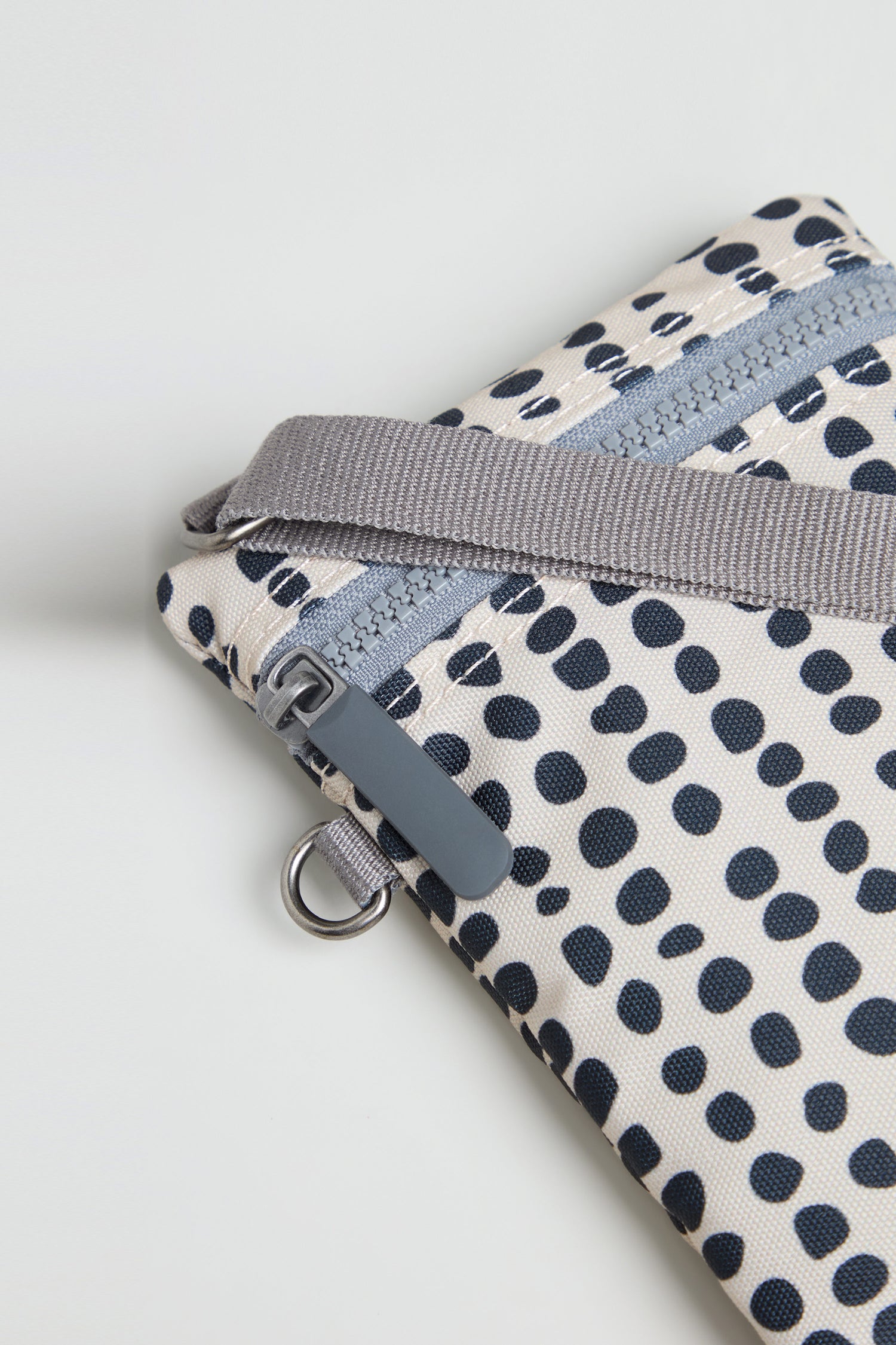 A close-up of the Recycled Canvas Chelsea Dotty Bag, showcasing navy polka dots, a gray zipper, and a gray strap attached to a metal loop.
