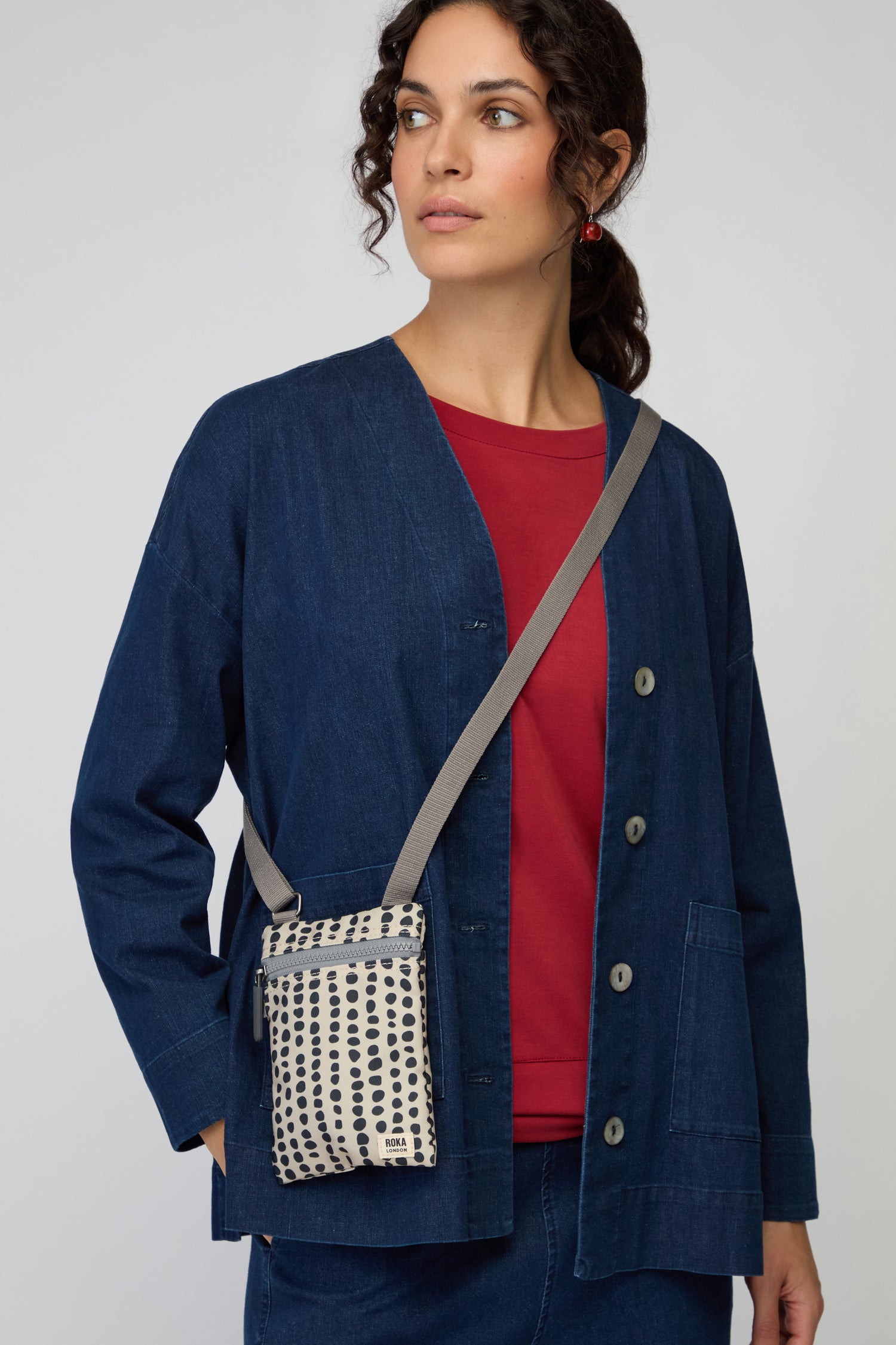 A woman in a blue buttoned jacket over a red top and blue jeans holds the Recycled Canvas Chelsea Dotty Bag—an elegant white crossbody with a black dotted pattern, crafted from recycled canvas.