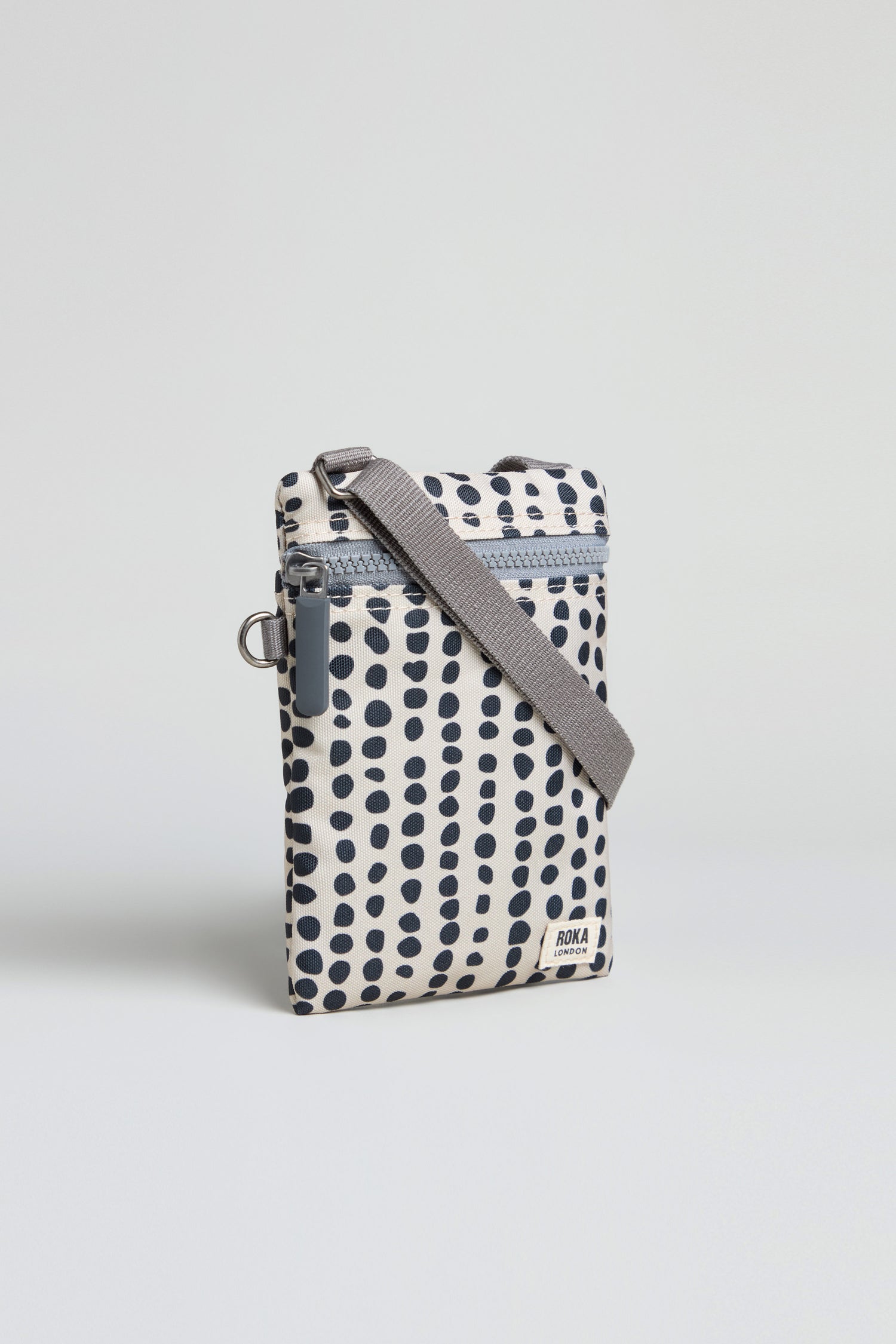 The Recycled Canvas Chelsea Dotty Bag is a small, rectangular crossbody with a gray strap and white background, featuring evenly spaced dark blue dots.