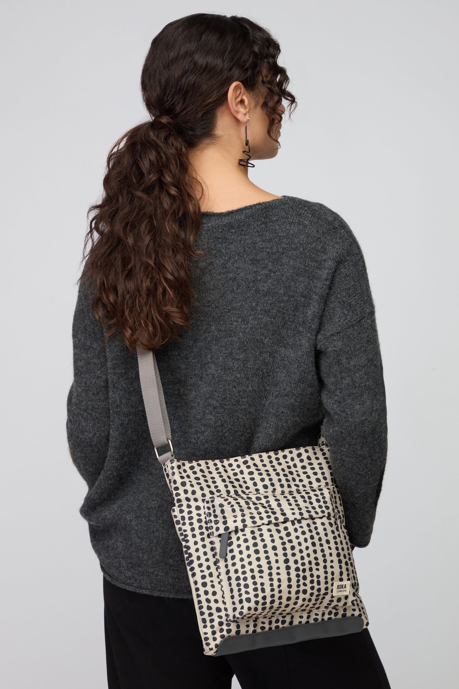 A woman with curly brown hair in a ponytail wears a dark gray sweater and black pants, carrying the Recycled Canvas Kennington Dotty Bag featuring a white and black dotted pattern.