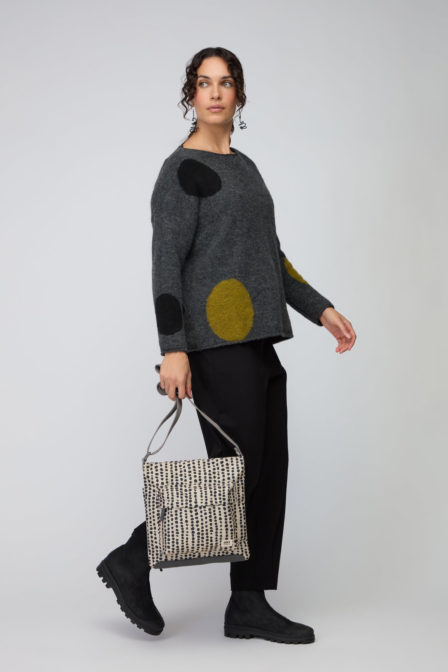 Wearing a gray sweater with large black and yellow circles, black pants, and black boots, a person carries the Recycled Canvas Kennington Dotty Bag—a weather-resistant crossbody—for both style and practicality.
