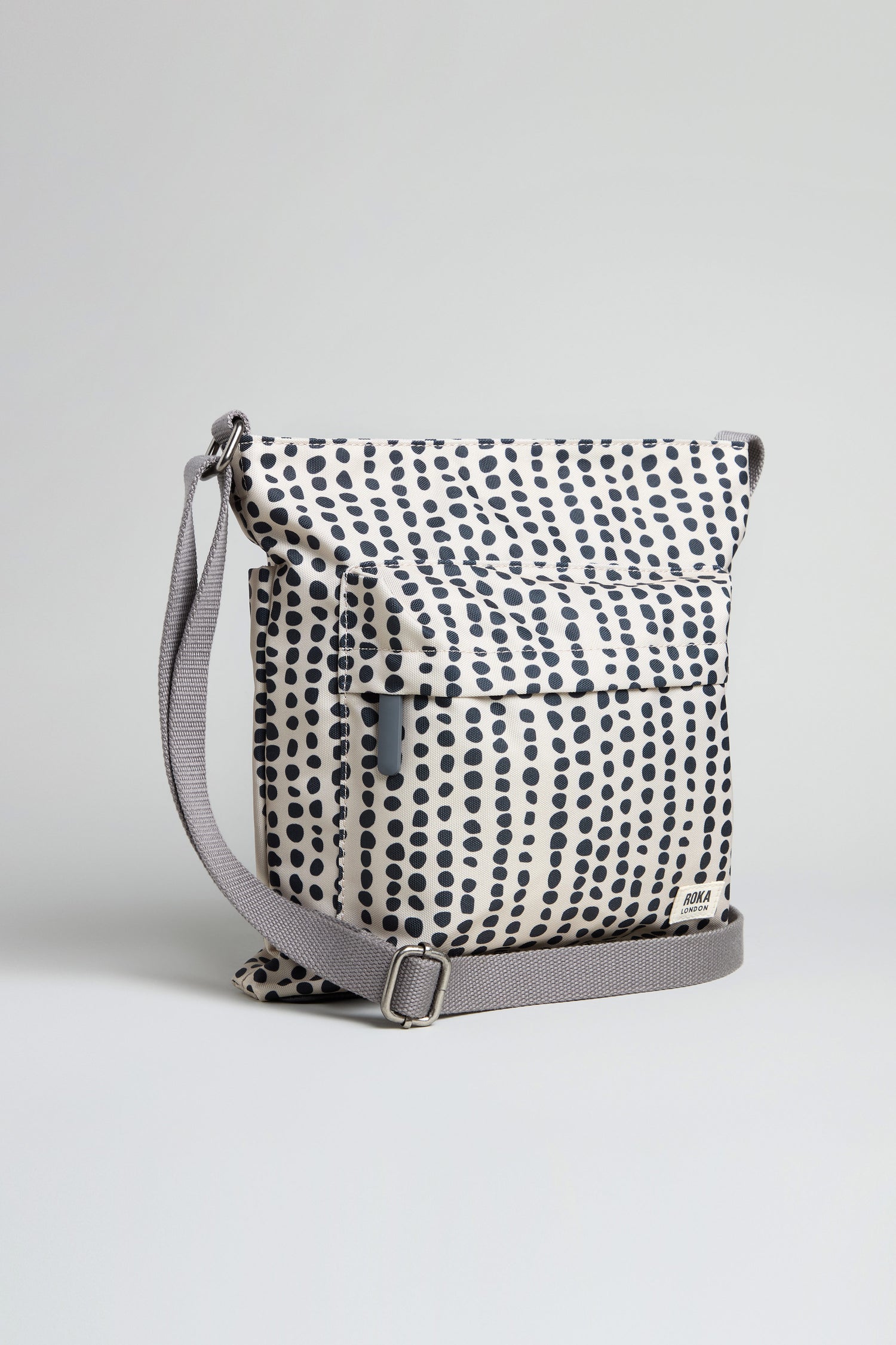 The Recycled Canvas Kennington Dotty Bag features a white base with black polka dots, a front pocket, flap closure, and an adjustable gray strap, displayed against a plain background.