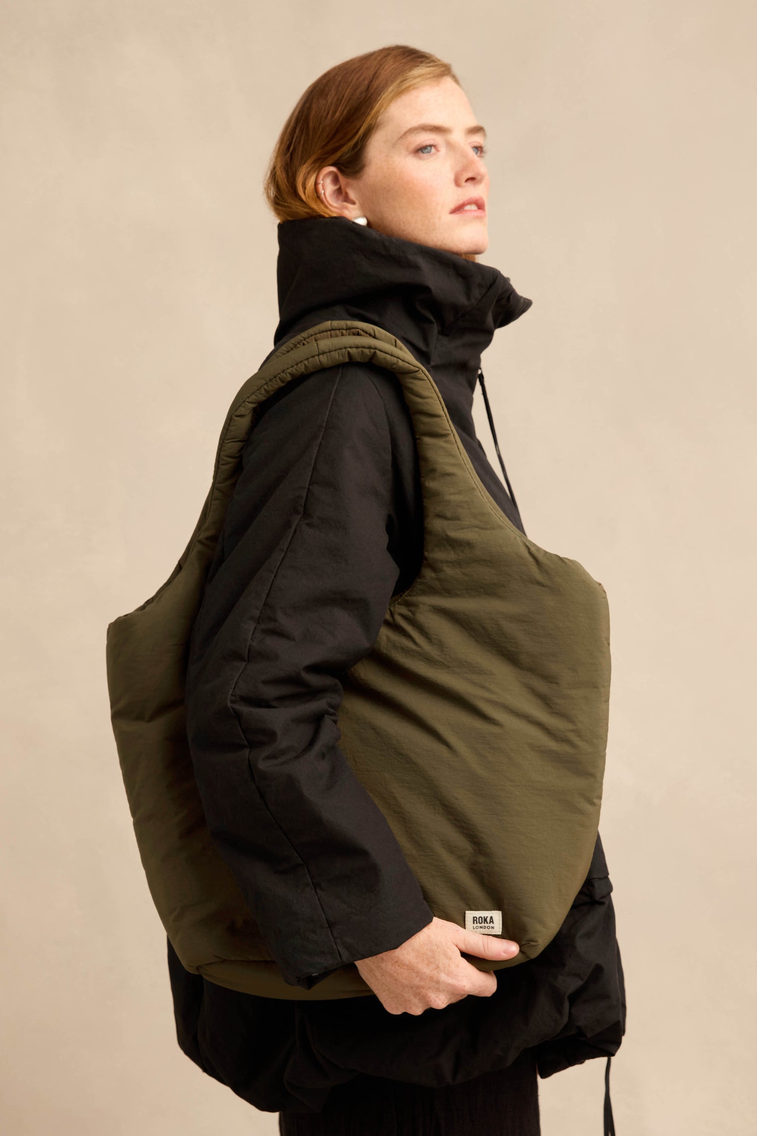 A person wearing a black jacket and carrying an oversized Oxford Moss Shoulder Bag—with an olive-green hue and a padded laptop sleeve—over their shoulder, standing against a plain beige background.