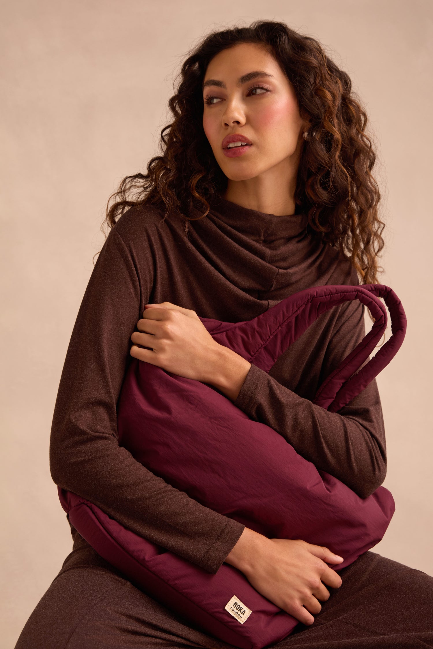 A woman with long curly hair, dressed in a brown outfit, sits holding the Oxford Merlot Shoulder Bag—a large maroon quilted bag featuring a padded laptop sleeve—against a plain beige background.