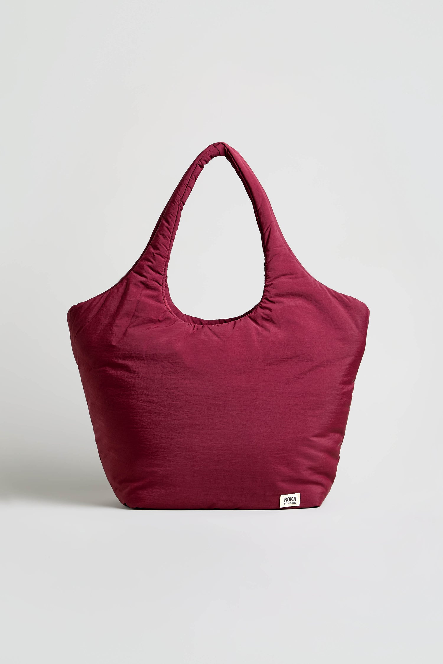 The Oxford Merlot Shoulder Bag is a large maroon fabric bag with wide handles, a small white brand label at the bottom, and a padded laptop sleeve for extra protection, displayed against a plain light background.