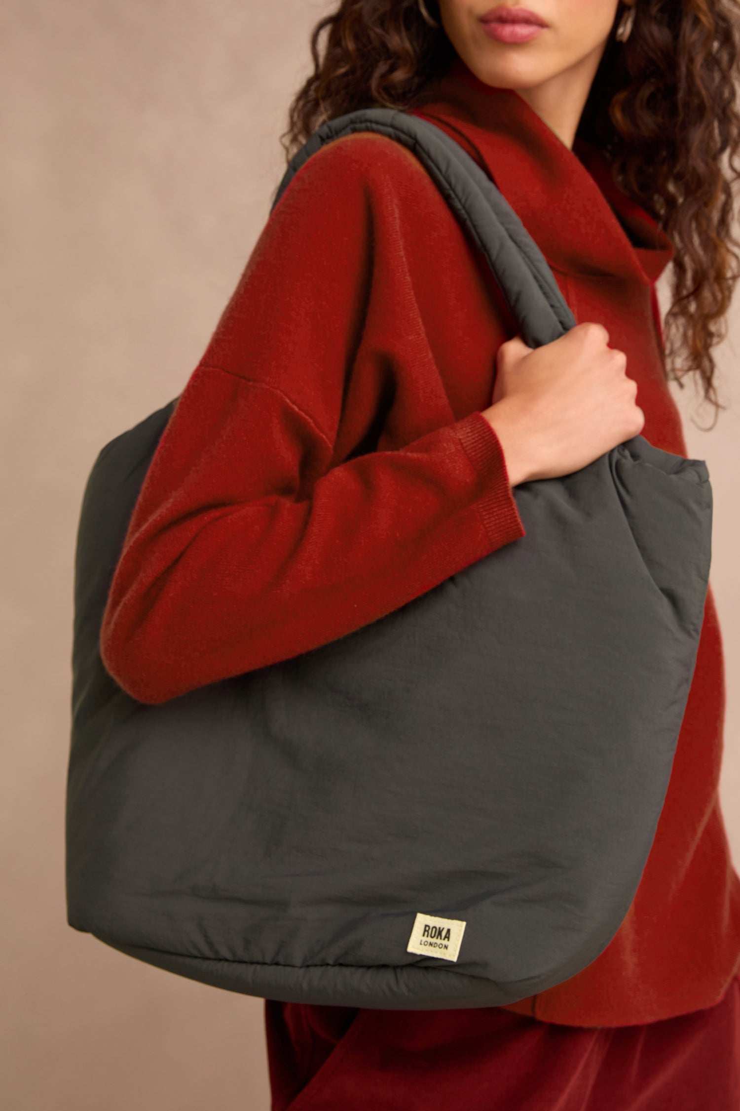 A person wearing a red sweater is holding a large, dark gray Oxford Raven Shoulder Bag with a visible “Roka” label on the lower corner. This weather-resistant shoulder bag features a padded laptop sleeve, blending style with practical functionality.