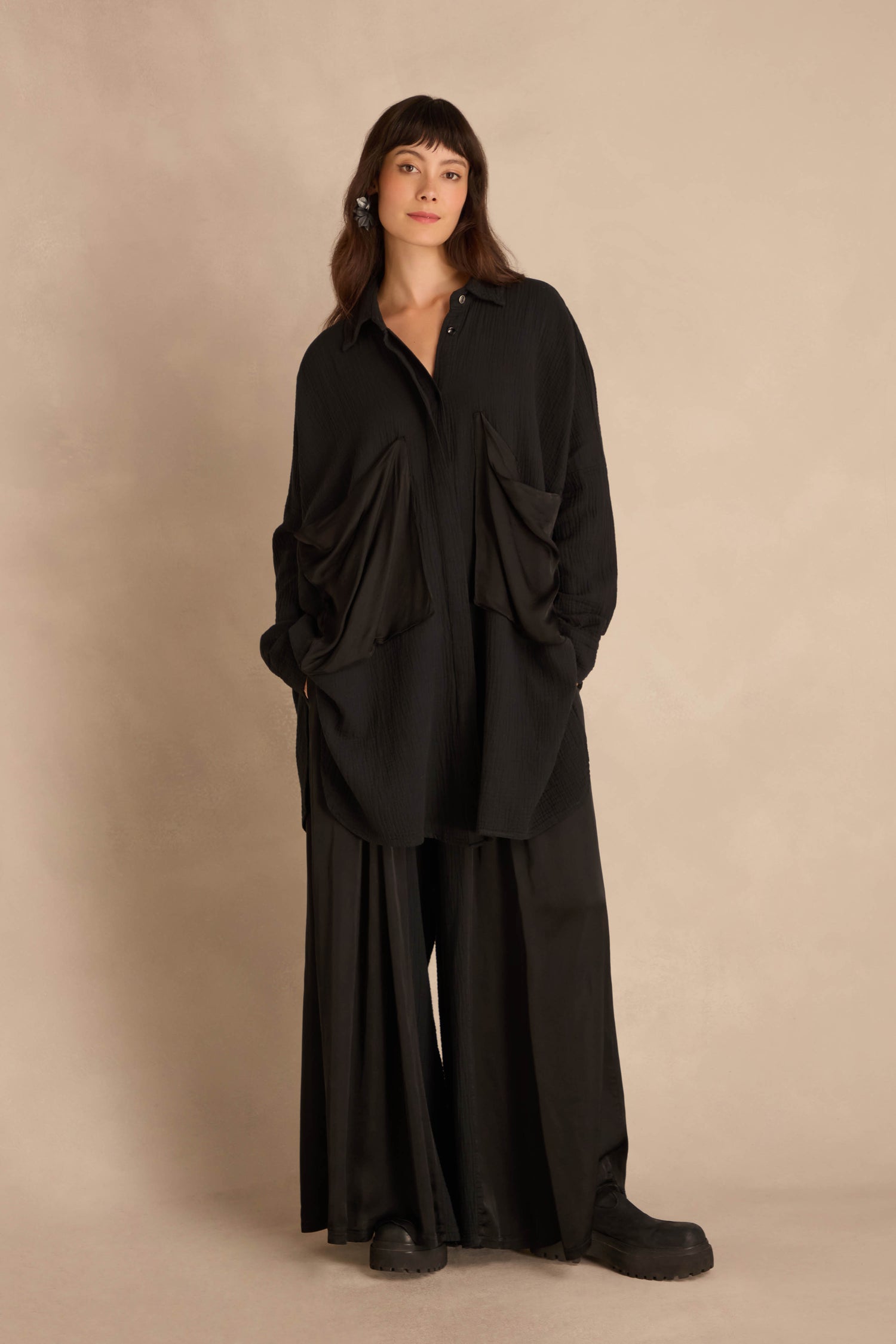A person wearing a Black Pocket Satin Shirt, featuring an oversized, crinkled cotton design with two satin patch pockets, paired with wide-leg trousers.