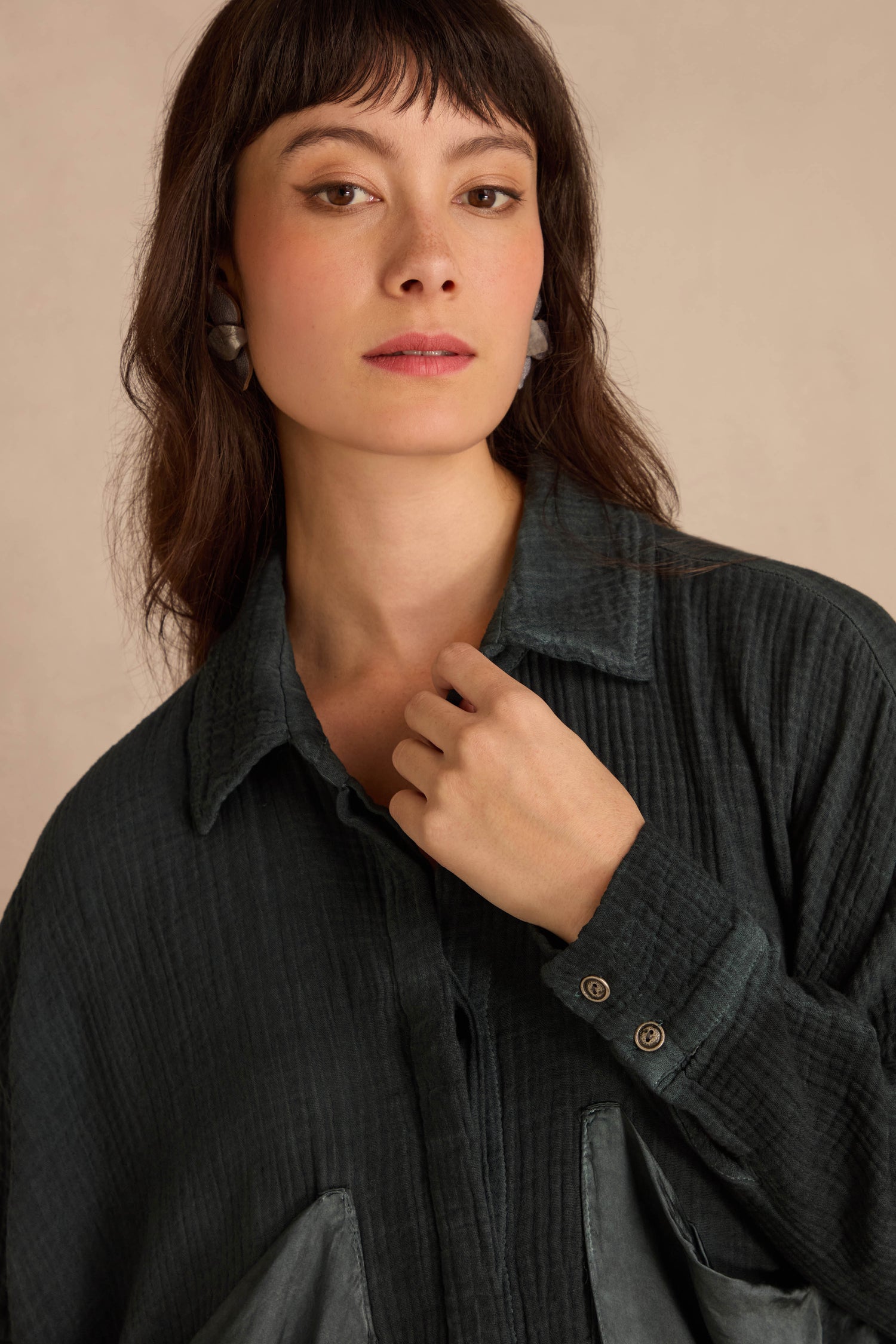 A person wearing an oversized, dark green, crinkled cotton Petrol Pocket Satin Shirt with a front button-down closure and pocket.