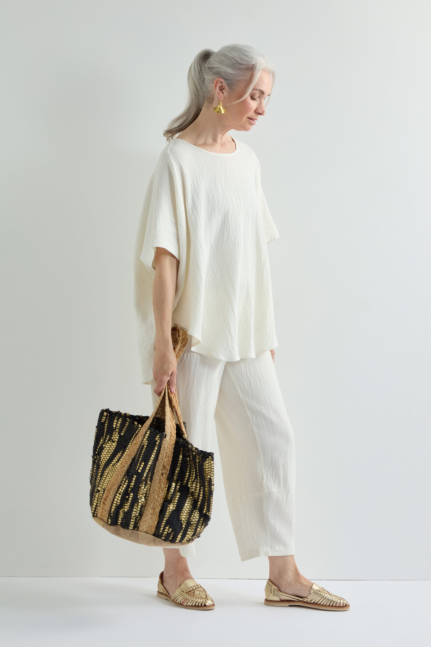 An older woman with gray hair in a ponytail wears a loose white top and pants, yellow earrings, gold shoes, and carries a Black and Gold Upcycled Leather Tote Bag, standing against a white background.
