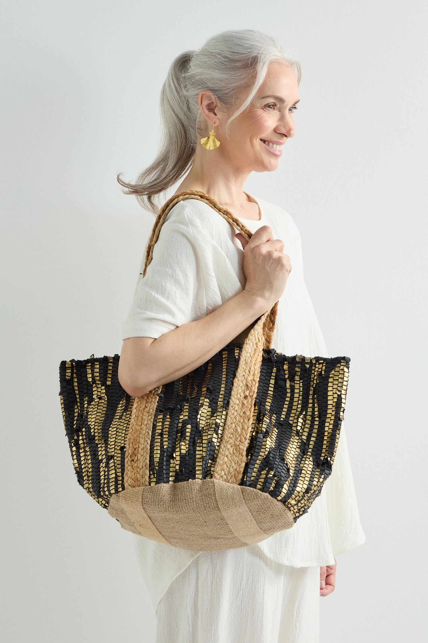 An older woman with gray hair in a ponytail, dressed in white and gold earrings, holds a Black and Gold Upcycled Leather Tote Bag handmade in India, featuring striped patterns that highlight sustainable craftsmanship.