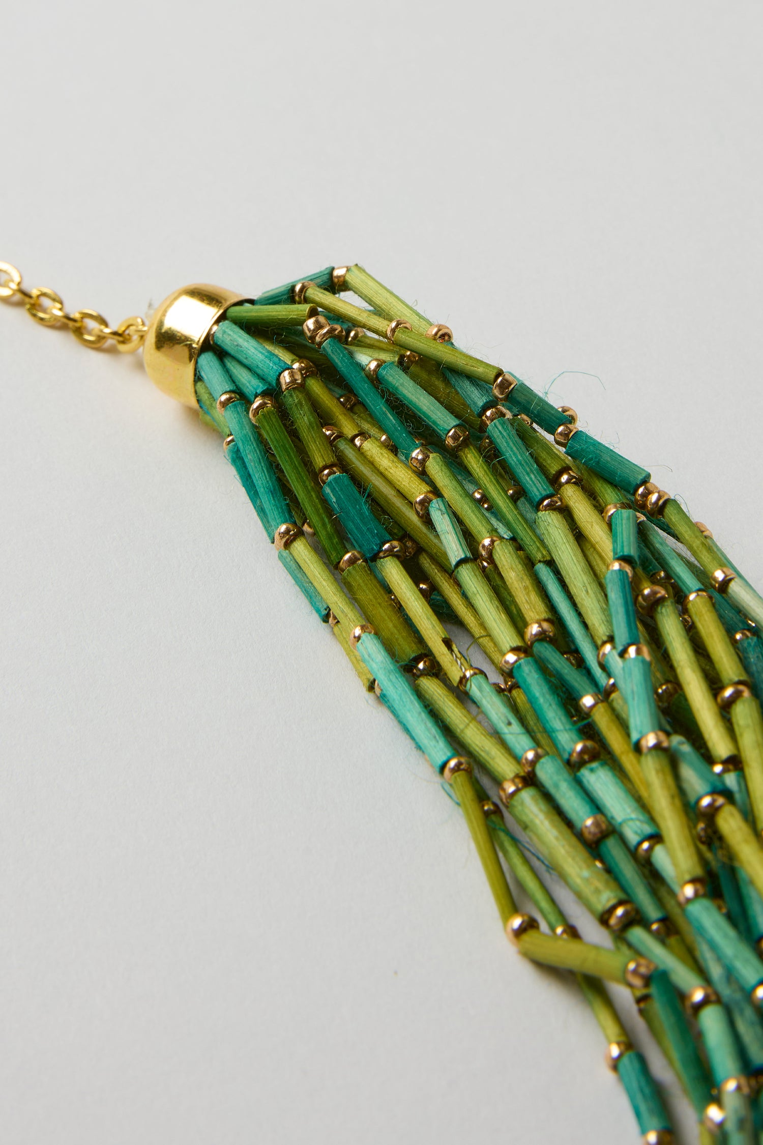 Close-up of the Multi Strand Grass Bead Necklace, featuring green and yellow cylindrical grass beads accented with small gold beads, a gold cap, and a gold-look clasp chain.