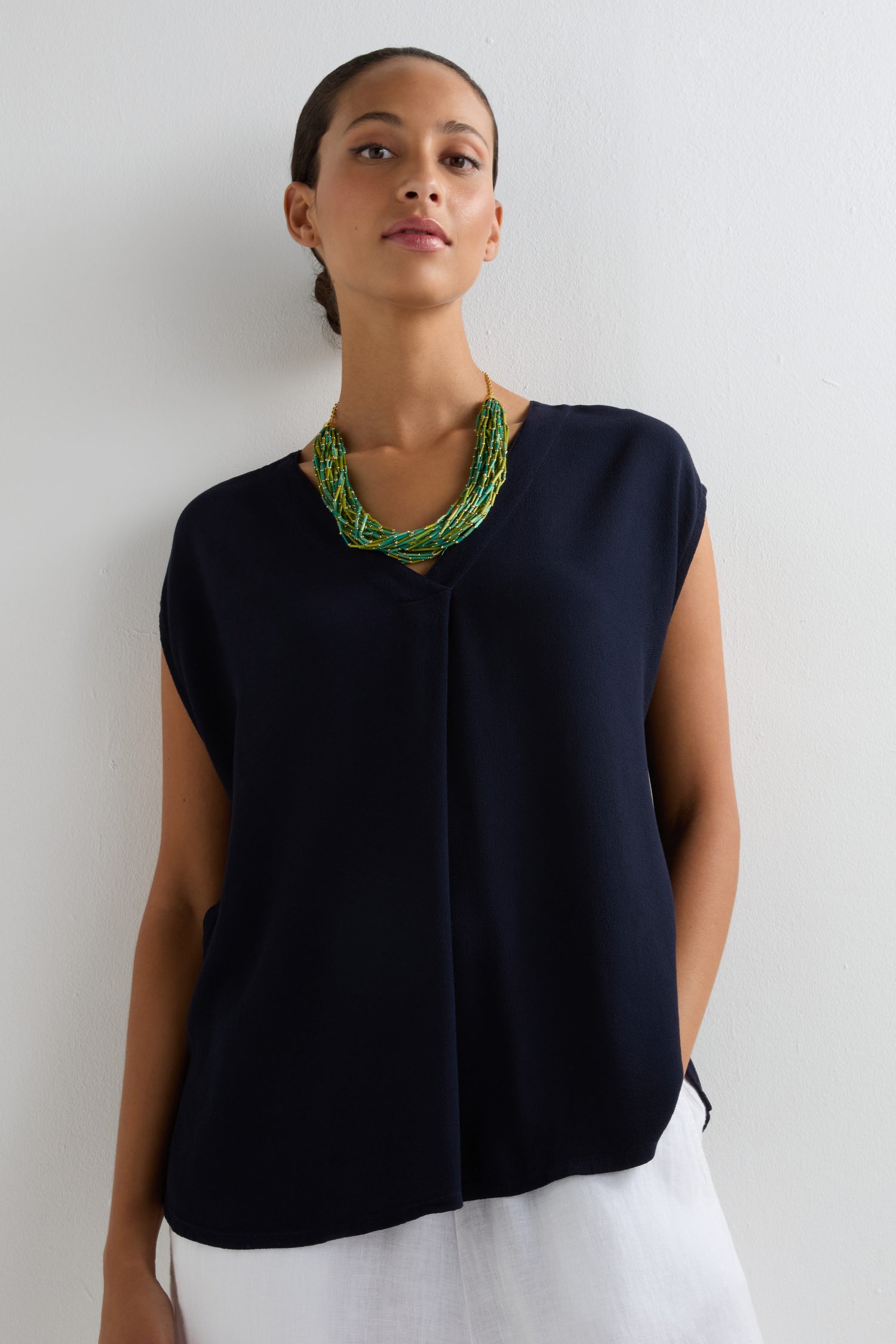 A person stands against a white wall wearing a sleeveless navy top, white pants, and the Multi Strand Grass Bead Necklace with a polished gold-look clasp chain.