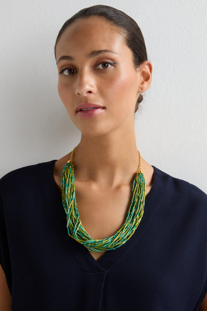 A woman with her hair pulled back wears a dark navy top and the Multi Strand Grass Bead Necklace, featuring vibrant grass beads and a gold-tone clasp, while standing against a plain white background.