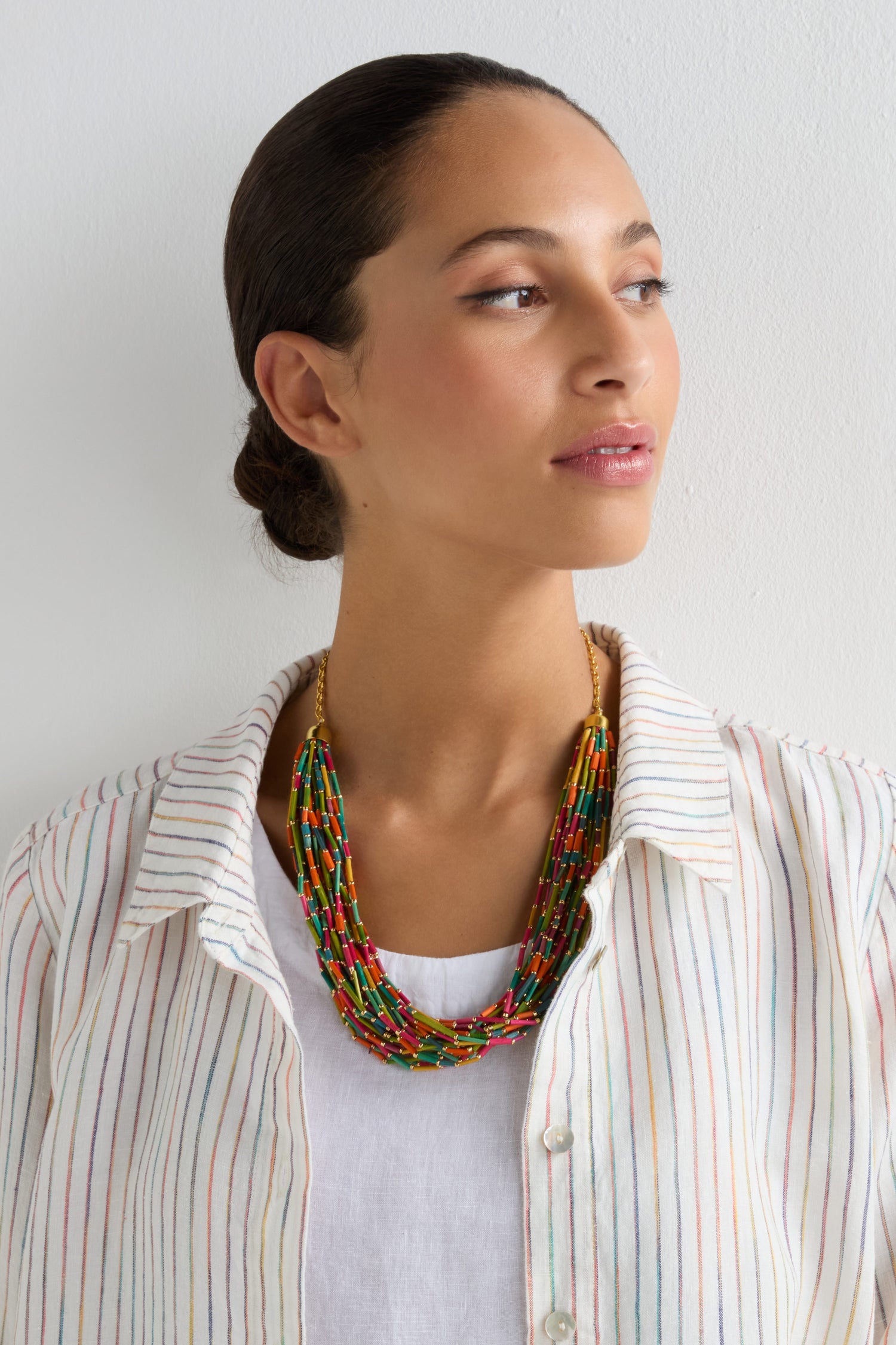 A woman with dark hair in a low bun wears a white shirt with colorful stripes and the Multi Strand Grass Bead Necklace, featuring vibrant grass beads, as she looks left against a plain white background.