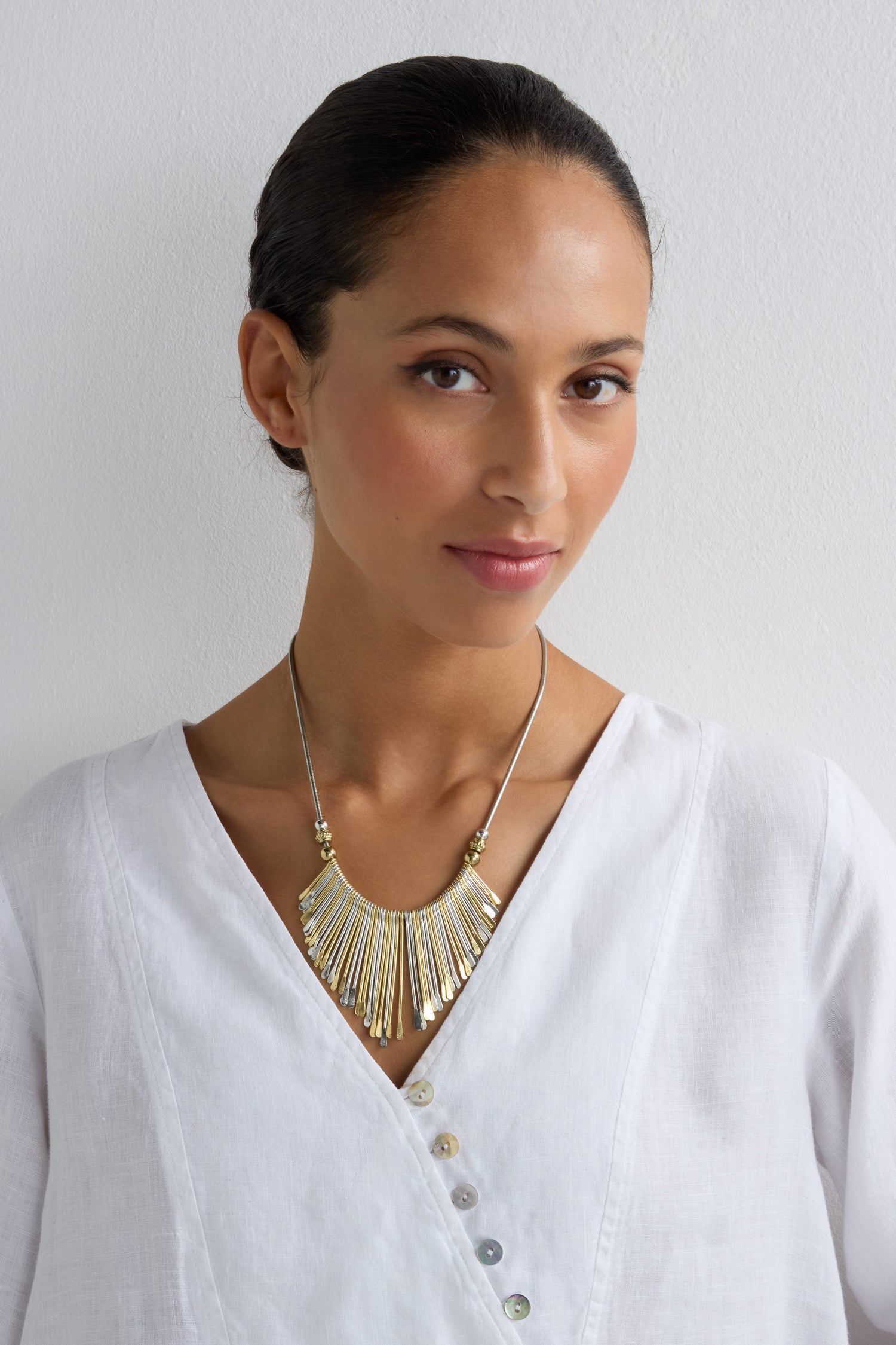 A woman wearing a white V-neck blouse and the Mixed Metal Fan Necklace poses against a plain white background, looking at the camera.