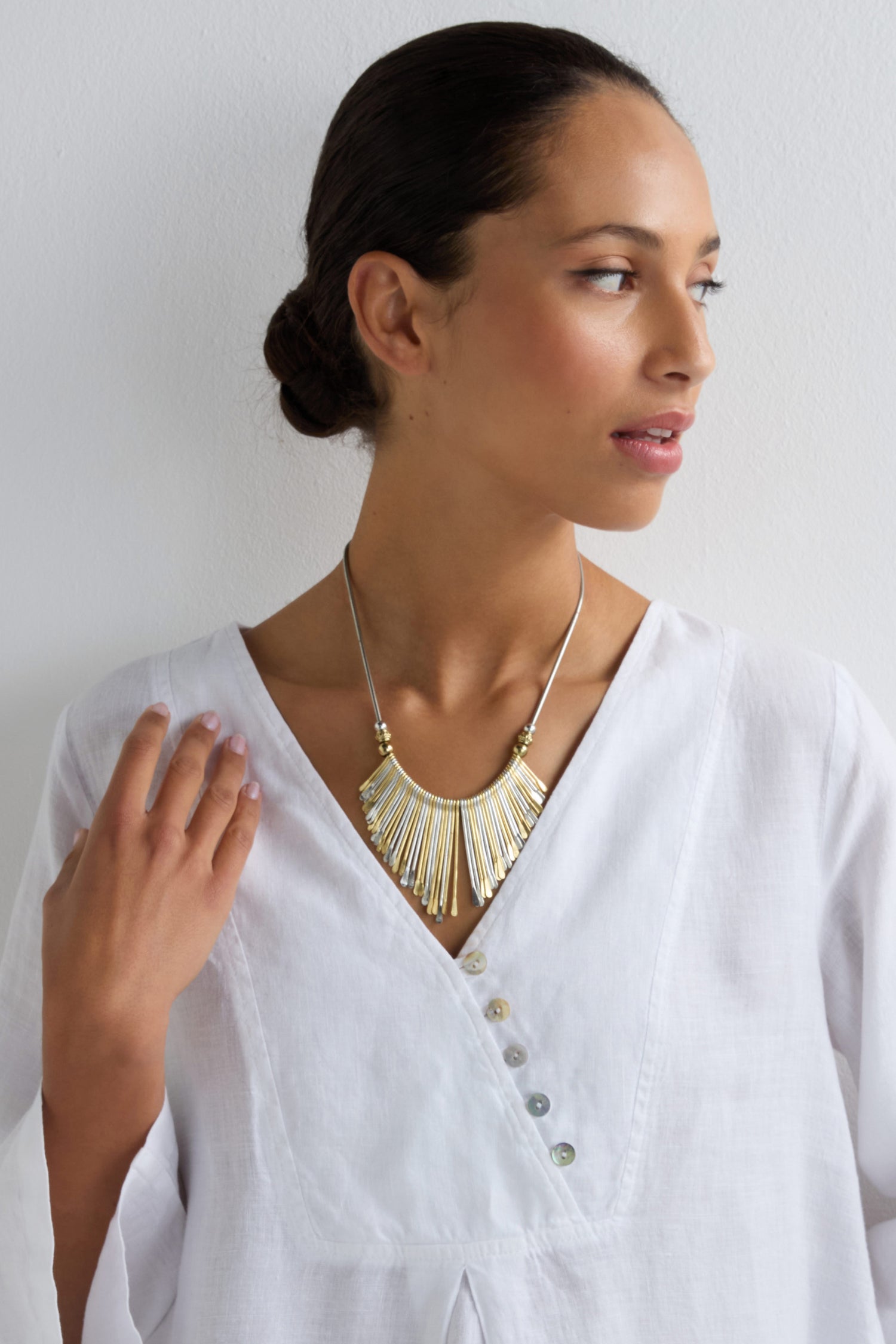 A woman in a white blouse gazes sideways, showcasing the Mixed Metal Fan Necklace, which features gold and silver-toned metalwork with fringe details, all set against a plain white background.