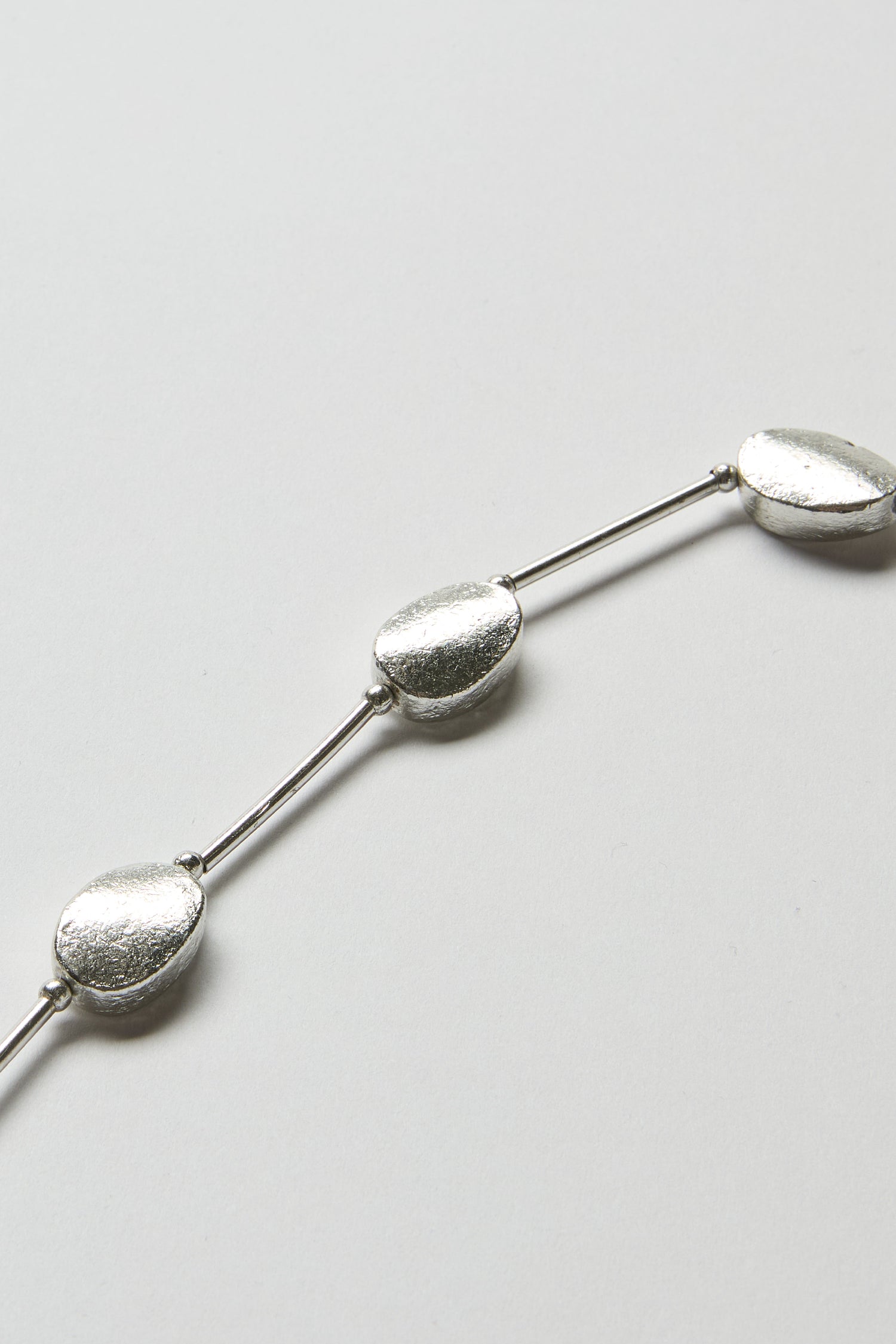 The Oval Silver Disc Necklace features oval-shaped silver beads linked by sleek connectors, offering quiet elegance and a modern design, showcased against a crisp white background.