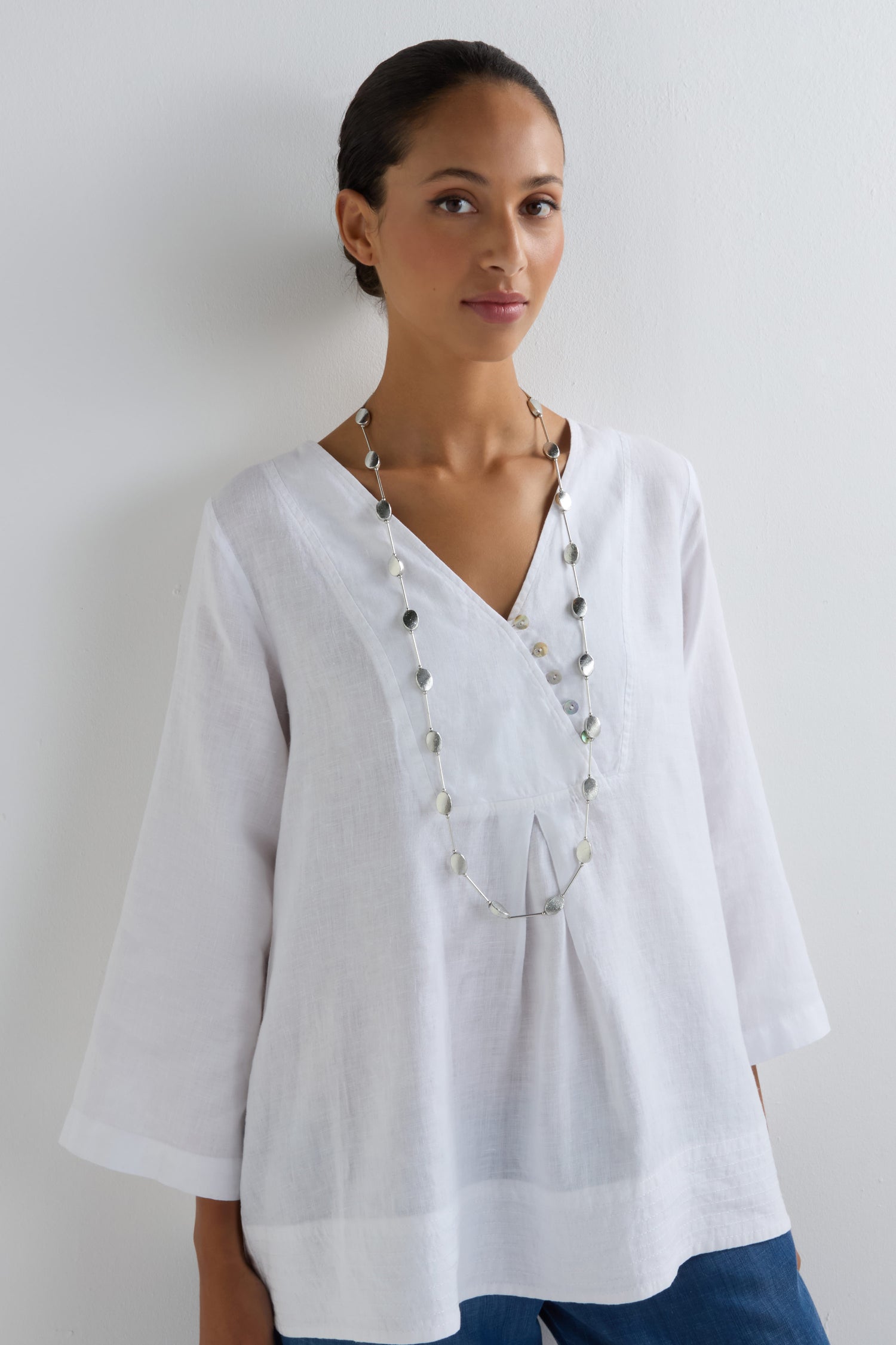 A woman in a white V-neck blouse poses against a plain white wall, showcasing the Oval Silver Disc Necklace.