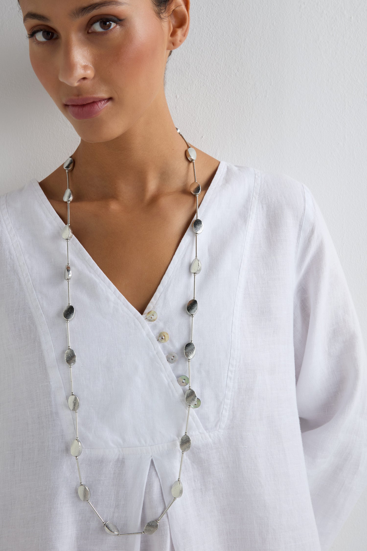 A woman stands against a plain white background, wearing a white V-neck blouse and the Oval Silver Disc Necklace.