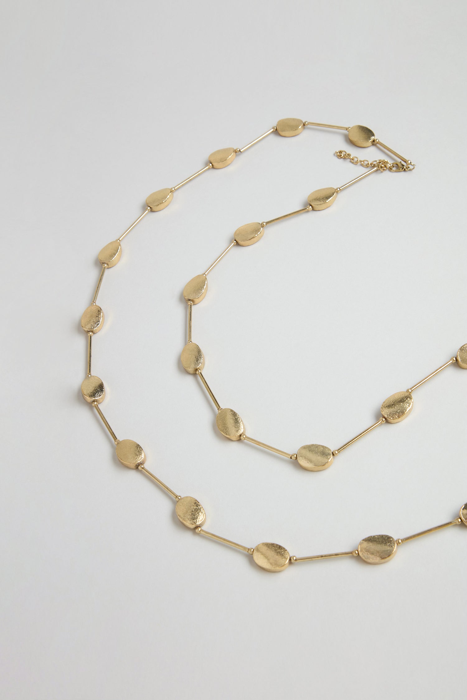 The Long Oval Disc Necklace features textured gold oval discs connected by short bars, displayed in a spiral arrangement on a plain white background.